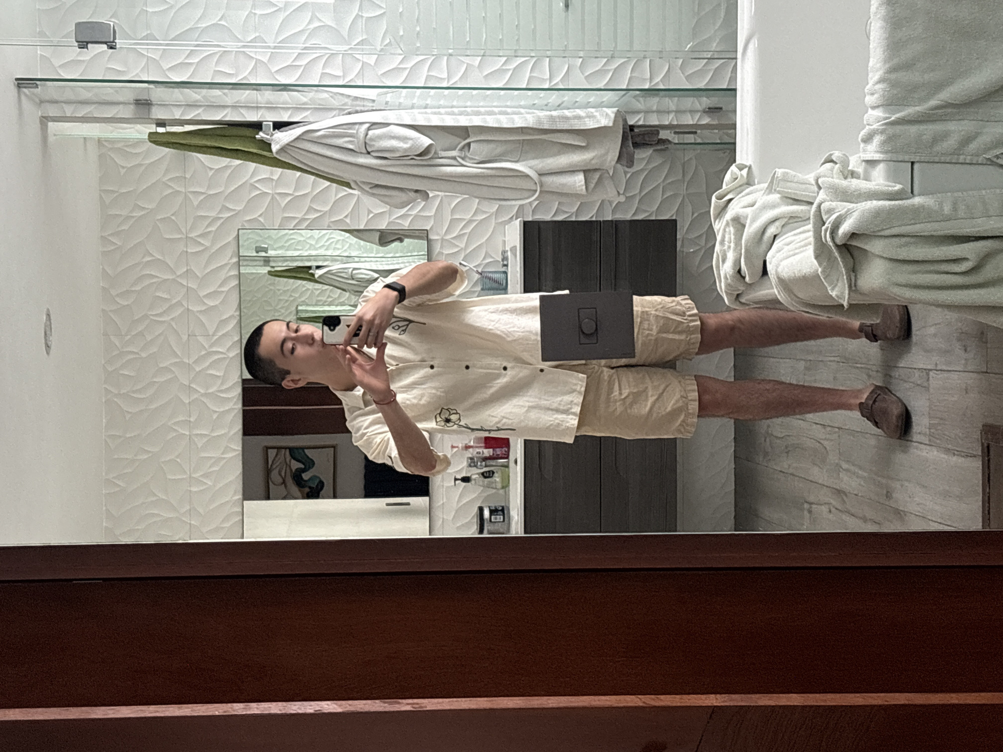 A person is taking a selfie in a bathroom. The person is a light-skinned male with short, dark hair. He is wearing a light-colored shirt with floral details and khaki shorts. He has a watch on his left wrist and a bracelet on his right. He is holding a cell phone up to his face in his right hand.
The bathroom has white walls with a textured leaf pattern. There is a glass shelf above the sink with folded white towels and a green towel. The vanity is dark gray with a mirror above it. There are toiletries on the counter. A stack of white towels is on a surface to the right. The floor is tiled in a light gray wood-grain pattern. The bottom of the image shows a dark wooden frame.
The location is implied to be in San Bernardino Tlaxcalancingo, Mexico.