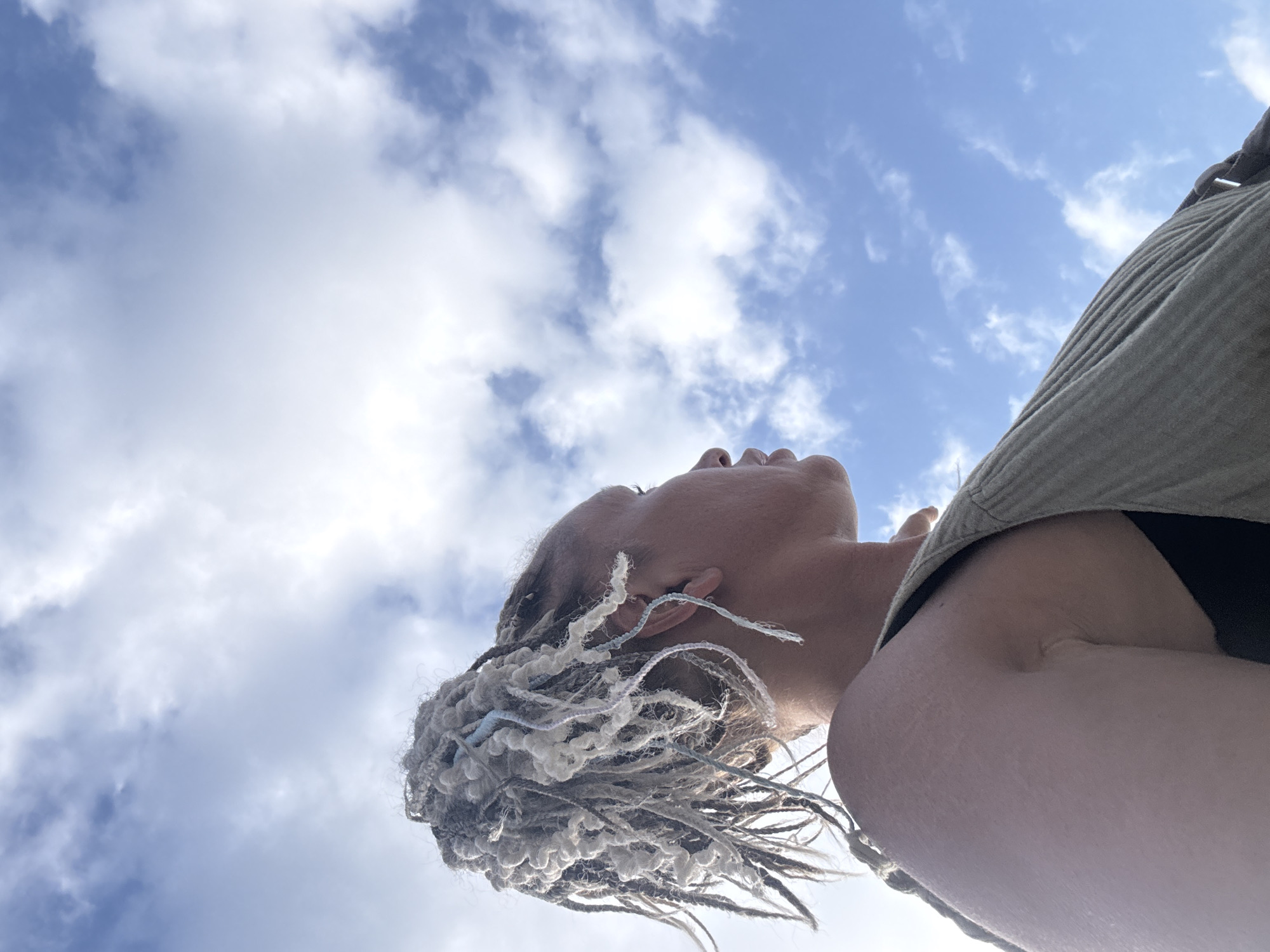 The photograph is taken from a low angle, looking up at a person with braided hair against a partly cloudy sky. The person's head is tilted back, with their face visible in profile. They are wearing a light-colored shirt or jacket. The background consists of a blue sky with white, fluffy clouds. No specific details indicate that this image was taken in Kuta Selatan, Indonesia. More information is required to determine location.