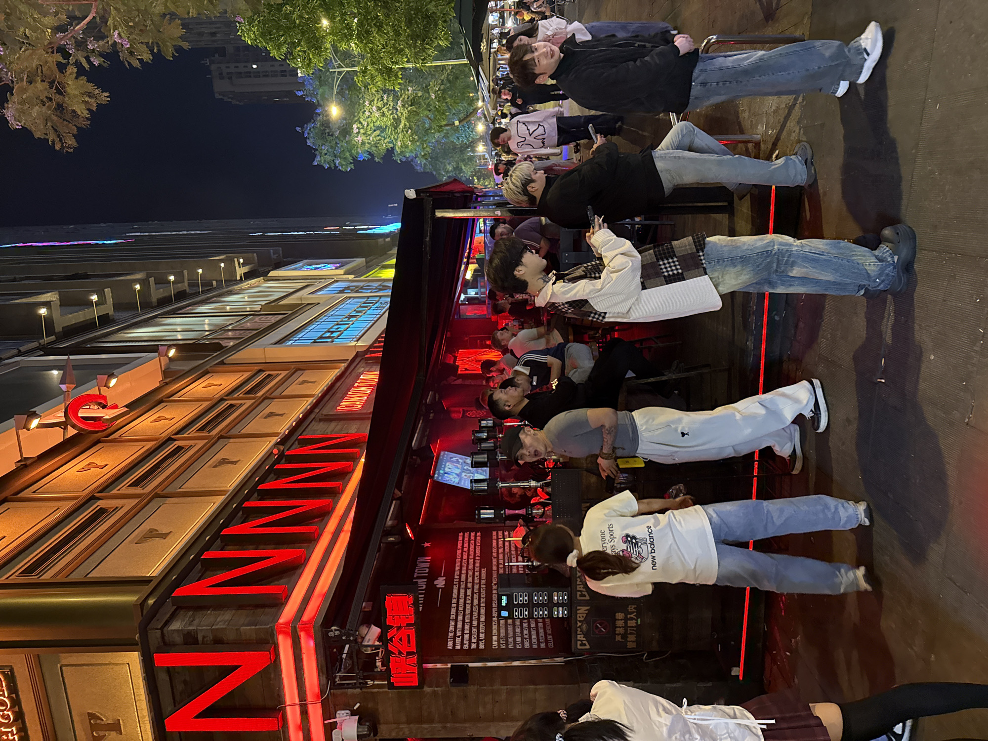 Here's a matter-of-fact description of the image:
The image depicts an outdoor nighttime scene in what appears to be a bustling entertainment area, possibly the city of Liangjiang, China. The main focus is a bar named "Canyon Town". The exterior of the bar is decorated with large, illuminated red signage in English.
Several people are seated at tables outside the bar, engaging in activities such as talking, using their mobile phones, or simply observing the surroundings. The individuals appear to be diverse in age and attire.
The environment includes a building with illuminated windows and awnings. Above, there are trees and streetlights. The overall ambiance is lively and urban.