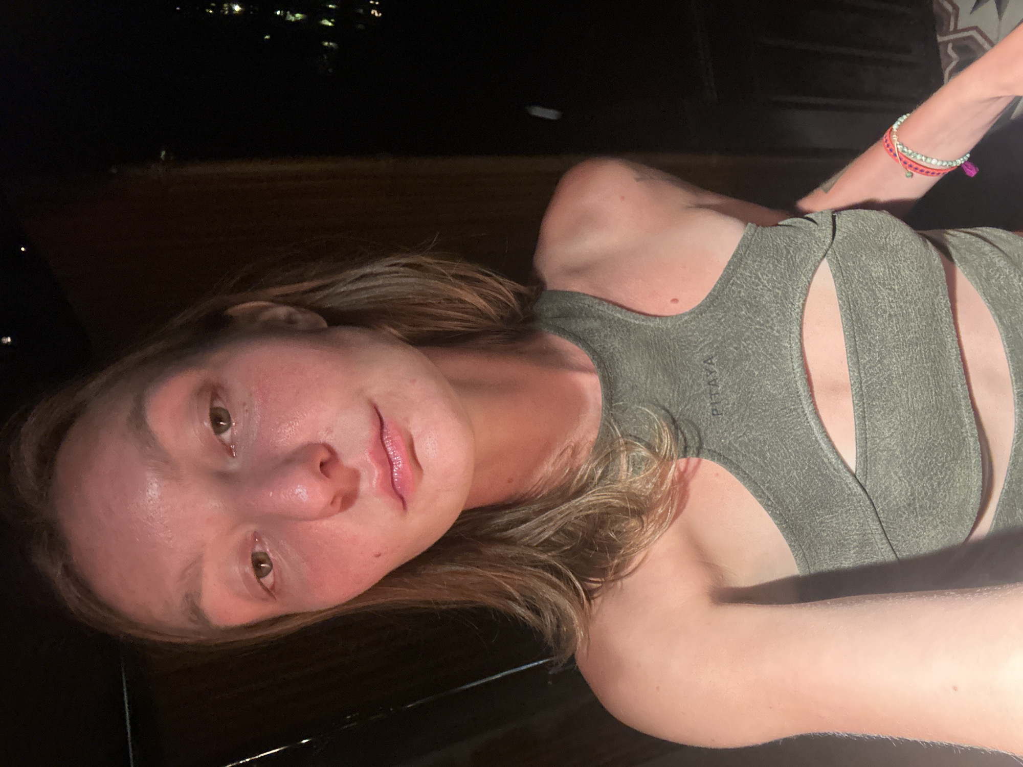 A young adult female is depicted in a close-up, chest-up selfie, facing directly towards the camera. She has fair skin with a reddish tone, possibly from lighting, and dark eyes gazing directly at the camera with a neutral expression. Her lips are closed, slightly parted, and appear glossy. Her medium-length, light brown hair frames her face.
She wears a dark olive green, textured athletic top with a high neck and a horizontal cut-out design across the chest. The brand 'PITAYA' is visible on the left shoulder strap. On her right wrist, she has a multi-layered bracelet with pink, green, and white beads and woven elements.
The background immediately behind her on the left is dark wood paneling. To her right and in the distant background, the scene is black with small, scattered points of light, indicating an outdoor nighttime environment. The illumination is dim, casting a warm glow on the subject's face and upper body. This scene is set in Mù Cang Chải, Vietnam.
