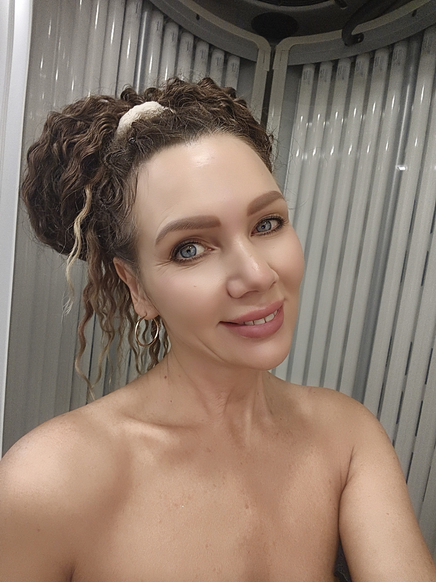 A woman with curly brown hair pulled up in a bun and large hoop earrings is shown from the chest up, smiling at the camera. She is in what appears to be a tanning bed with vertical, ribbed walls. Her skin is lightly tanned. The lighting is bright and even.