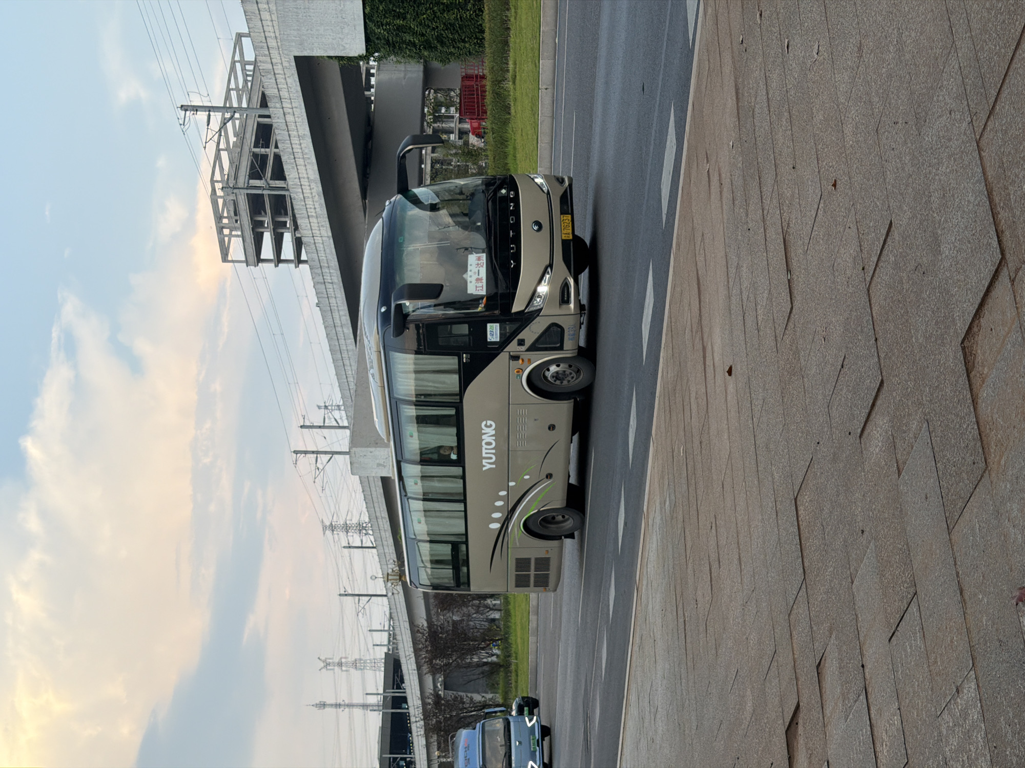 The image shows a beige Yutong bus driving on a road in Jiangjin, China. The bus has "YUTONG" written in white on its side. The bus is driving in the right lane of a multi-lane road. There is a sidewalk in the foreground made of large rectangular stone pavers. In the background, there is an elevated structure, possibly an overpass or part of an unfinished building, with electrical poles and wires visible. The sky is cloudy with a soft, diffused light, suggesting it may be late afternoon or early morning. A person can be seen through the bus window. Another smaller light-colored vehicle is visible to the left of the bus, also on the road. A red dumpster is visible in the far background to the right of the bus.