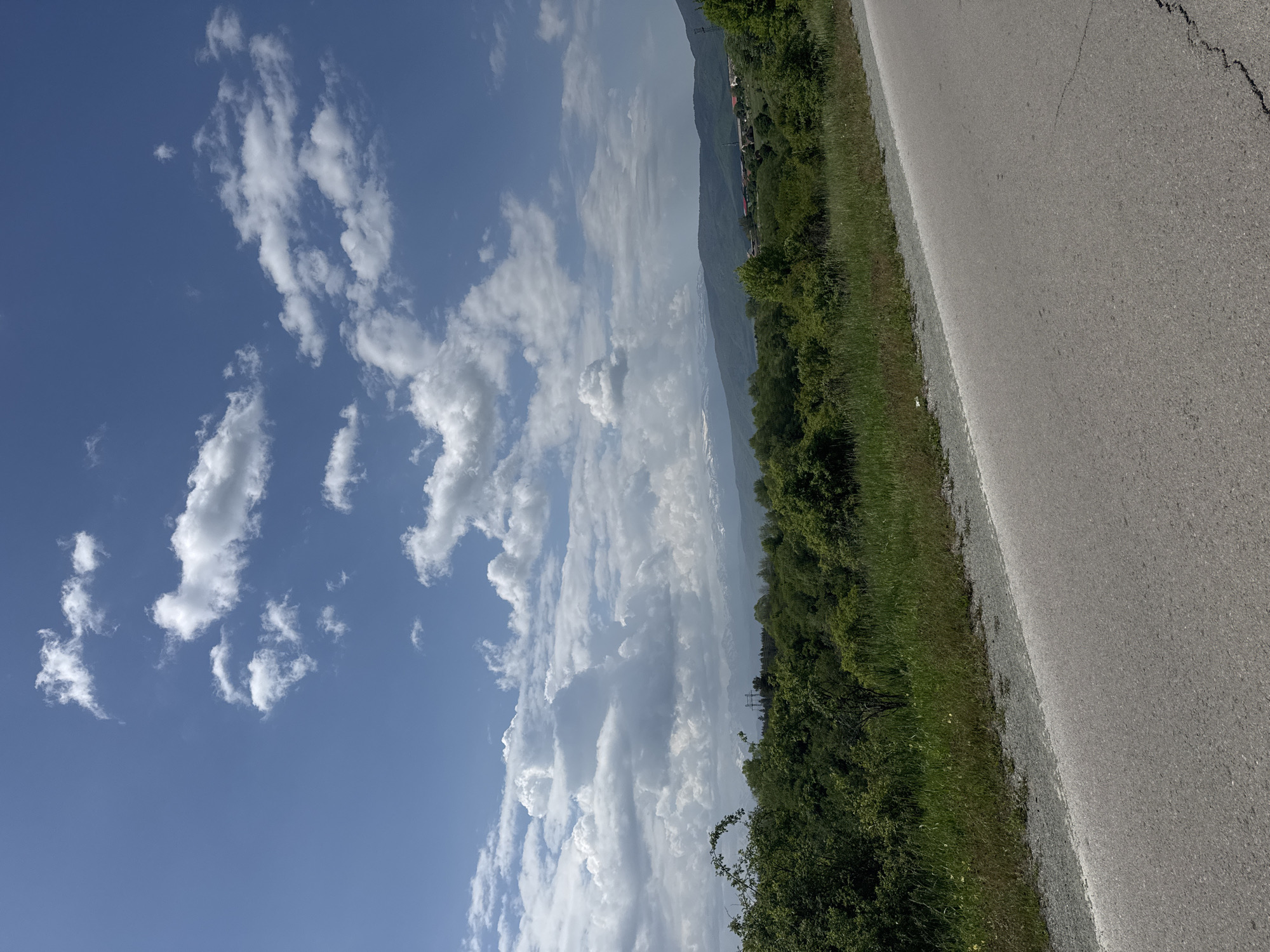 A photograph taken outdoors in Kovachevtsi, Bulgaria, shows a road stretching into the distance, parallel to vegetation. The sky is blue and contains cumulus clouds. The greenery consists of grass and shrubs. In the distance, mountains are visible. The road is asphalt. There are no people visible in the frame.