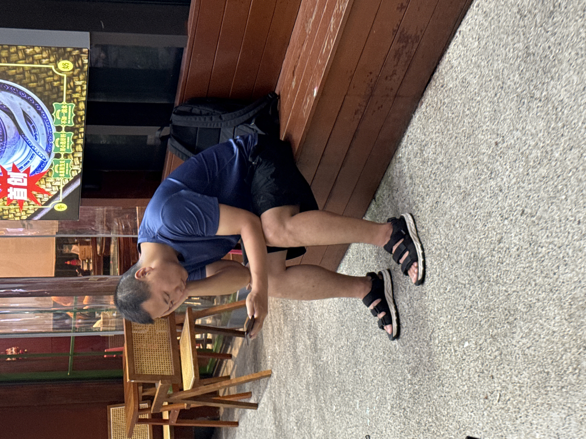 A man with short black hair and a blue t-shirt is sitting on a brown wooden bench, looking down at his phone. He is wearing black shorts and black sandals with white soles. His left arm is resting on his leg, and his head is propped up by his hand. A black backpack is behind him on the bench. In the background, there are tables and chairs, and a sign with Chinese characters. The ground is covered in a coarse, grey, speckled material. The setting appears to be an outdoor seating area of a restaurant or cafe in Yuzhong District, China.