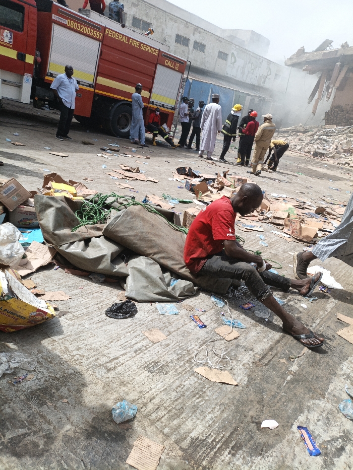 The image depicts the aftermath of an incident in Fagge C, Nigeria, characterized by structural damage and emergency response. In the foreground, a dark-skinned male in a red t-shirt and dark trousers sits on debris-strewn concrete ground, adjacent to a large olive-green tarp containing coiled green ropes. The ground is littered with cardboard pieces, plastic wrappers, and miscellaneous refuse.
In the midground, a red Federal Fire Service truck is parked, displaying "FEDERAL FIRE SERVICE" and "CALL 112". Several individuals are present around the truck and further right. These include civilians and emergency personnel in various attire: one male in a light blue shirt, another in a light-colored traditional garment, and uniformed responders wearing yellow helmets, red helmets, and khaki uniforms. One responder in a khaki uniform is observed bending down, interacting with ground-level debris.
The background features a multi-story light-colored building behind the fire truck. To its right, a significantly damaged building structure is visible, partially collapsed with exposed rebar, from which smoke or dust emanates, contributing to a hazy atmosphere. The sky is bright, indicating daylight conditions.