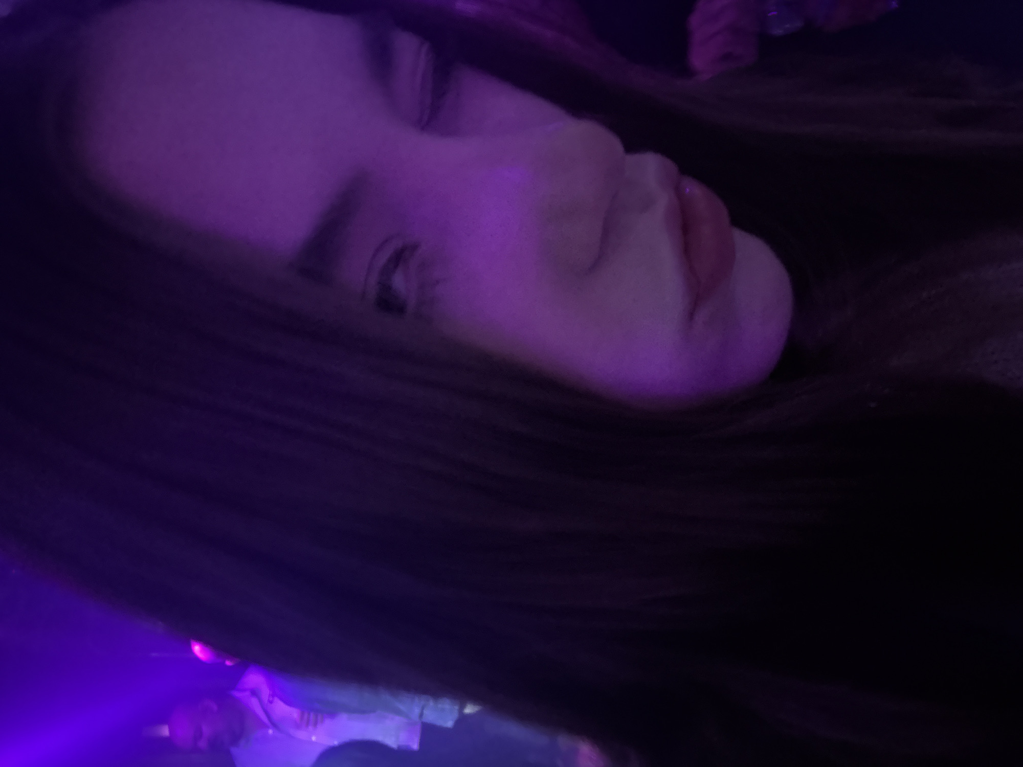 A close-up, canted angle portrait of a woman with long brown hair, illuminated by purple lighting. The background appears to be a dimly lit space with other people present but indistinct. Location cannot be determined from image.