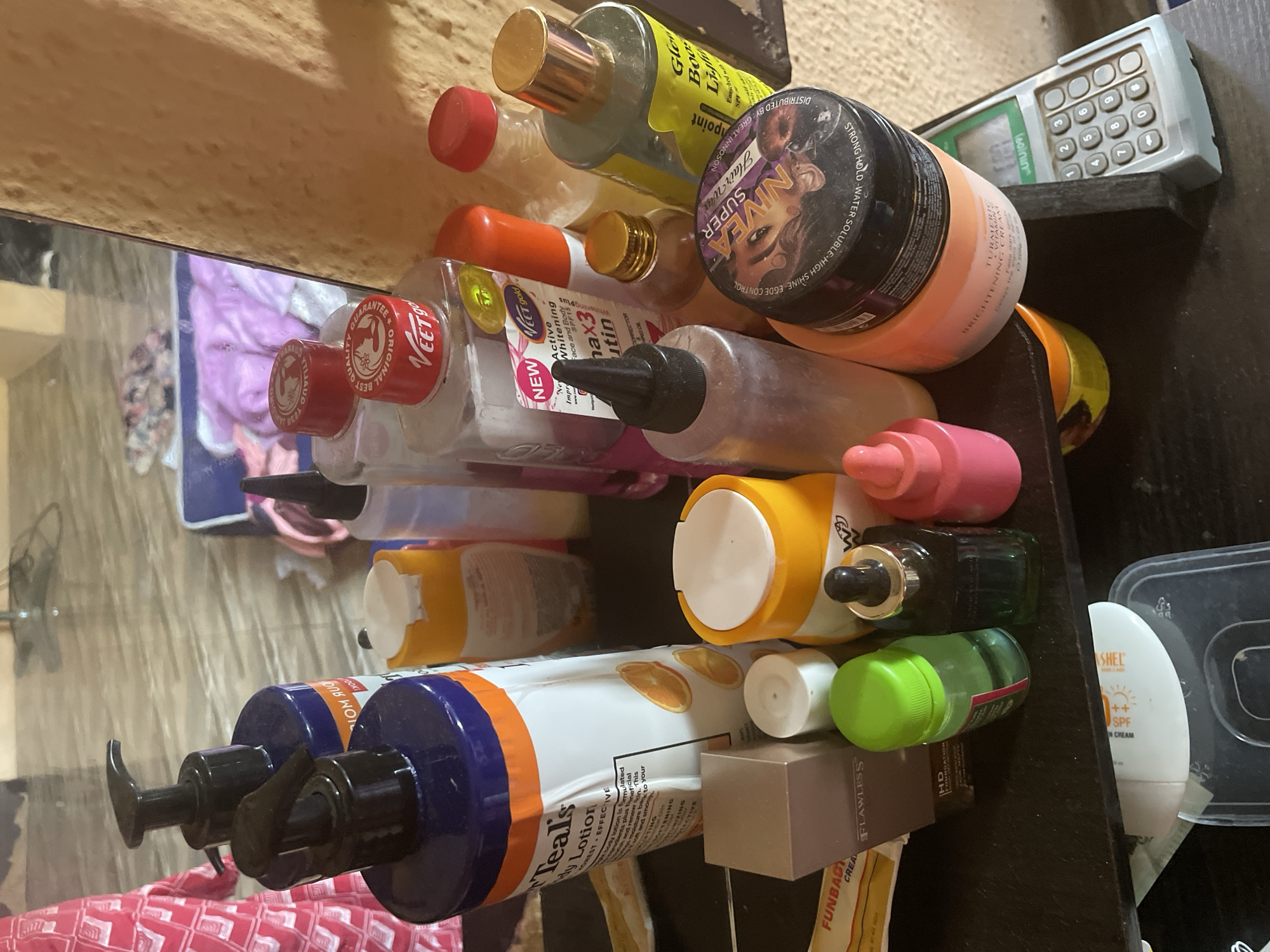 The image shows a shelf with various cosmetic products, including bottles of lotion, creams, and other skincare items, some of which are in a bathroom setting, possibly in Kakau, Nigeria. The products are arranged on a dark surface, and the background suggests a tiled floor and a wall. The lighting is dim, casting shadows. No people are visible in the image. The focus is on the collection of beauty products, implying a personal care or salon environment.