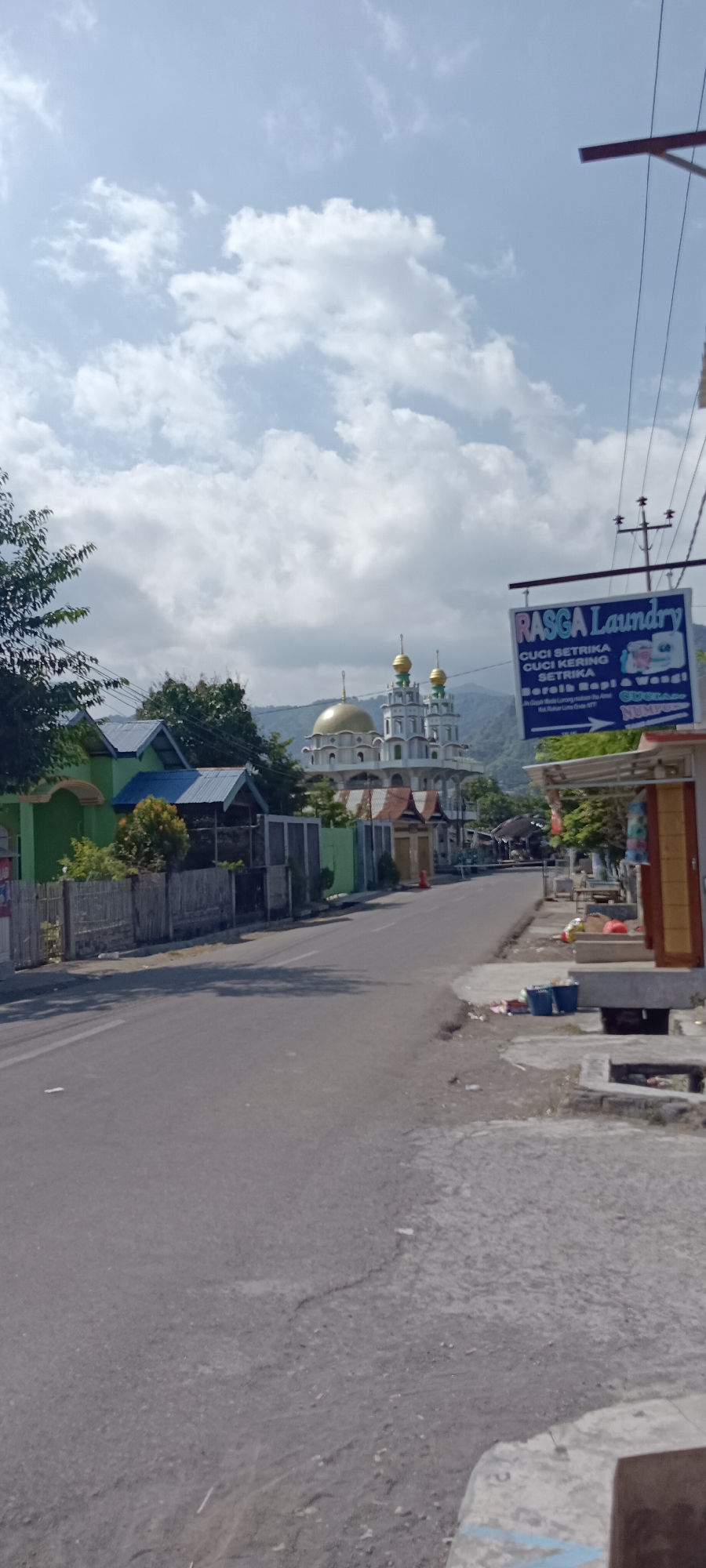 The image depicts a street scene in Ende, Indonesia, under a partly cloudy sky. On the left side of the road, there are several buildings, including one painted bright green with a blue-roofed structure attached. A bamboo fence runs along the property line. In the center of the image, a mosque with a prominent gold dome and two minarets stands tall, surrounded by other buildings. The road itself appears to be paved and shows signs of wear. On the right side of the road, a sign for "RASGA Laundry" is prominently displayed, hanging from a utility pole. Below the sign, there's a building with an awning, possibly a shop, with some items visible on display and a few blue buckets on the ground. The overall environment suggests a developing urban or semi-urban area in Ende.