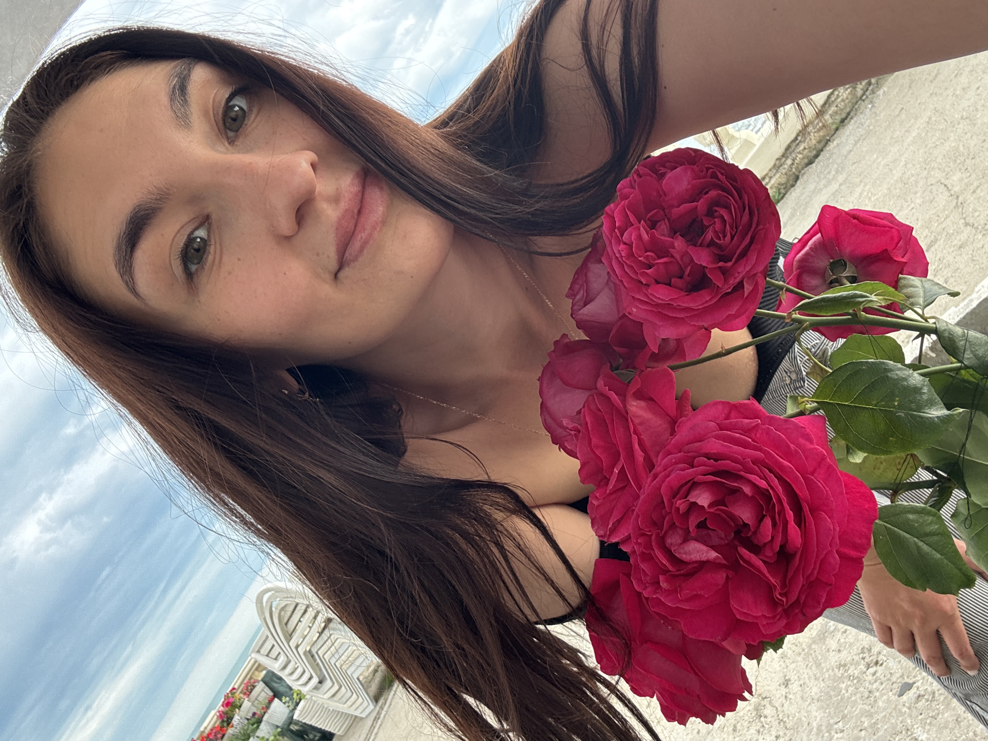 A selfie captures a young woman with long, dark brown hair and fair skin, holding a bouquet of deep red roses. Her gaze is directed at the camera, and she appears to be slightly smiling. A delicate gold necklace adorns her neck. She is dressed in black tank top with a button up shirt in black and white thin vertical lines, holding the roses in front of her.
The backdrop reveals a cityscape, suggestive of the city of Sochi, Russia. A white, ornate balcony railing can be seen in the background, overlooking what appears to be the Black Sea and a cloudy sky. Other features of Sochi are out of focus in the background.