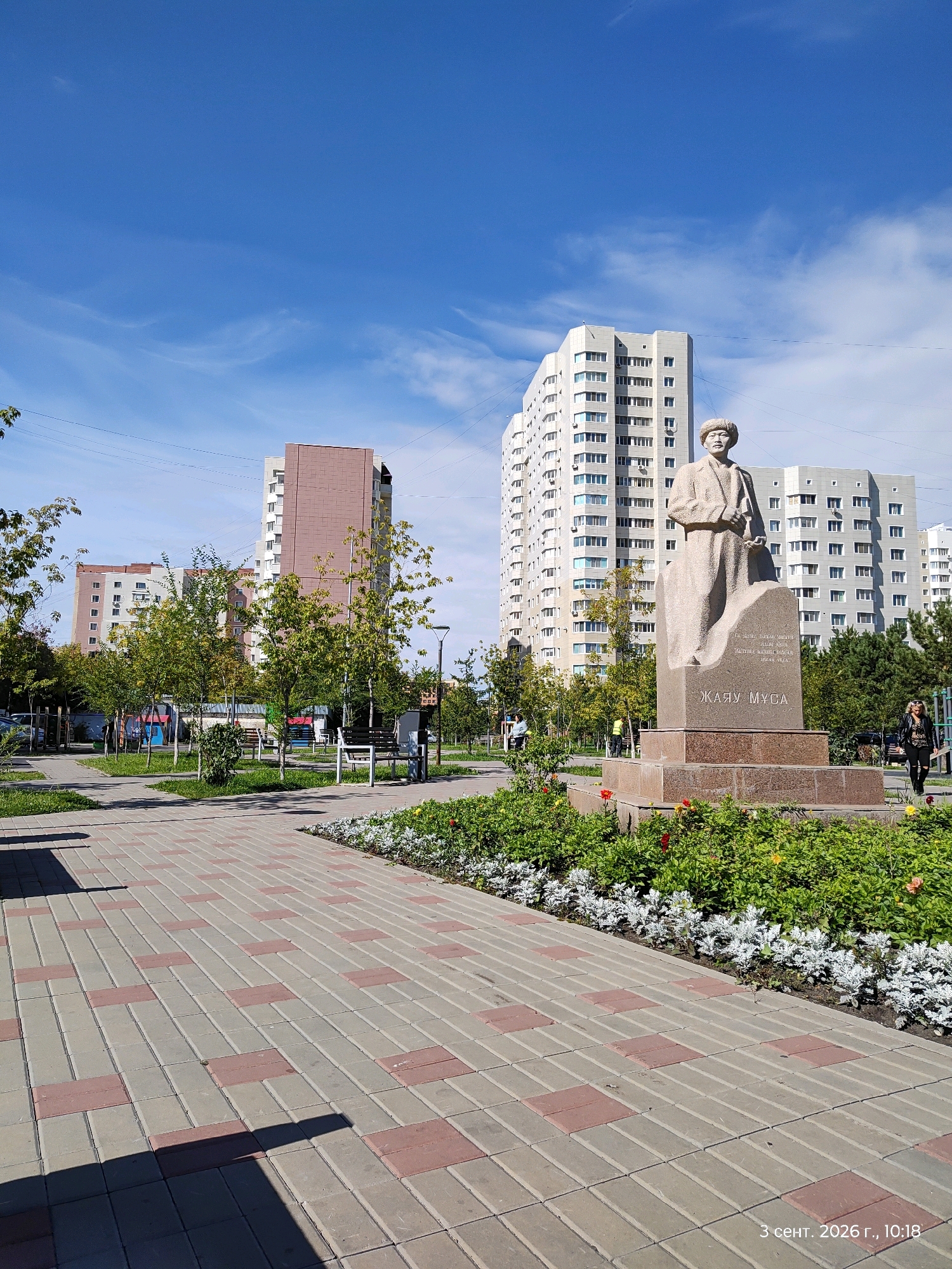 The image captures a sunlit urban scene with a monument at its center. The monument, made of stone, depicts a standing figure wearing a hat and coat, placed atop a granite pedestal inscribed with Cyrillic text. The pedestal is surrounded by a flowerbed with green foliage and red and yellow flowers. A paved walkway, laid with grey and red rectangular tiles, leads towards the monument.
In the background, modern multi-story apartment buildings with numerous windows dominate the skyline under a clear blue sky with wispy clouds. Several trees with green leaves are planted along the walkway, some of which are young saplings. Park benches are visible under the trees.
Two individuals are present in the scene: a woman in dark clothing walking to the right of the monument, and another person, partially obscured, near a bench on the left. A street light stands to the right of the monument. The overall environment suggests a public park or square within a city. The timestamp "3 сент. 2026 г., 10:18" is visible in the lower right corner. This is likely taken in Astana, Kazakhstan, given the monument's inscription which refers to Zhayu Musa, a prominent figure in Kazakh literature.