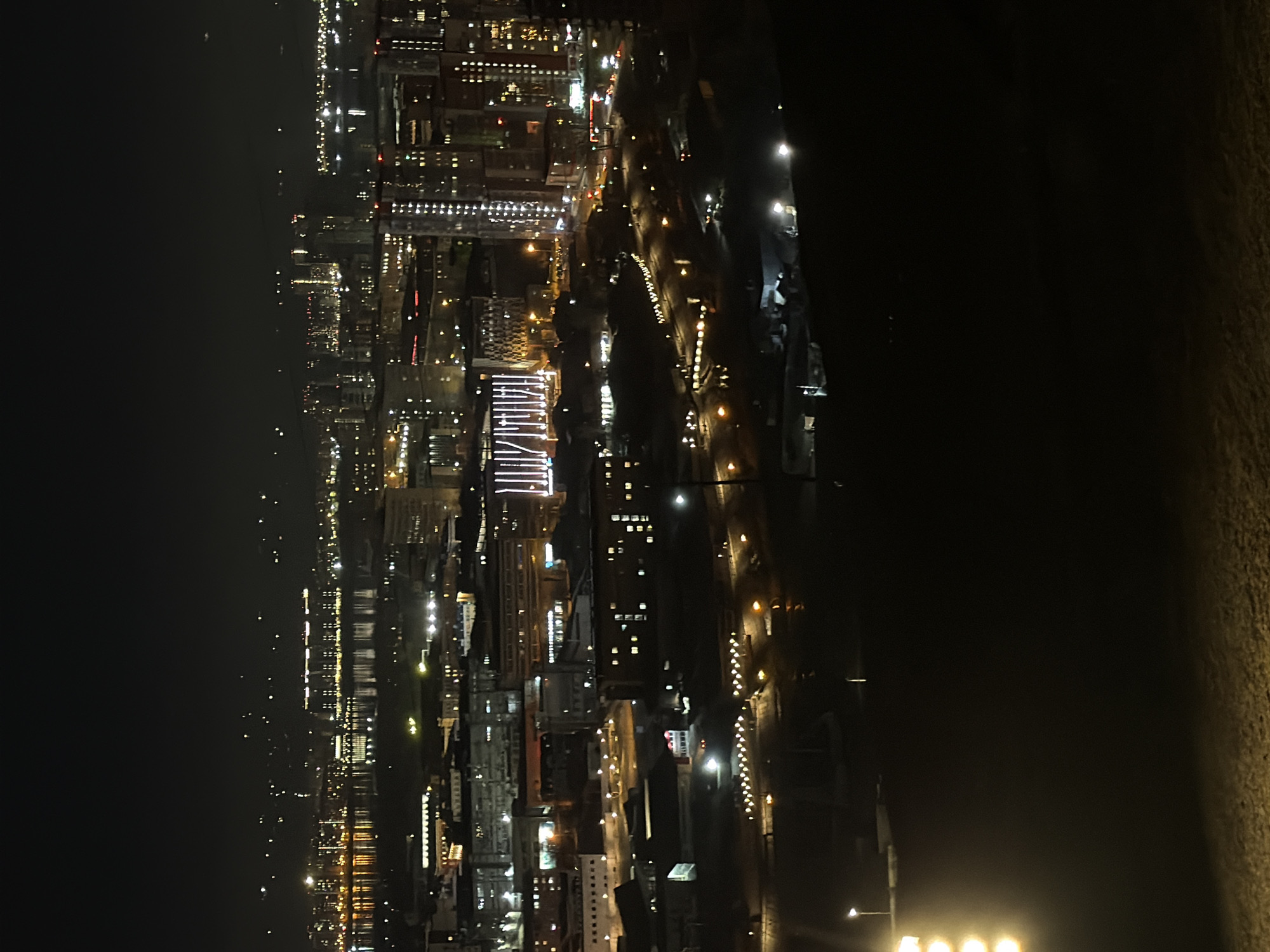 The image shows a nighttime view of Krasnoyarsk, Russia. The city skyline is illuminated with numerous lights from buildings, streets, and possibly boats on the Yenisey River. The lights vary in intensity and color, creating a complex pattern. The background is dark, suggesting it is a night scene and the viewer is likely at a high vantage point. To the right of the cityscape, there is a dark, undefined area, which is likely a natural feature like a hill or land.