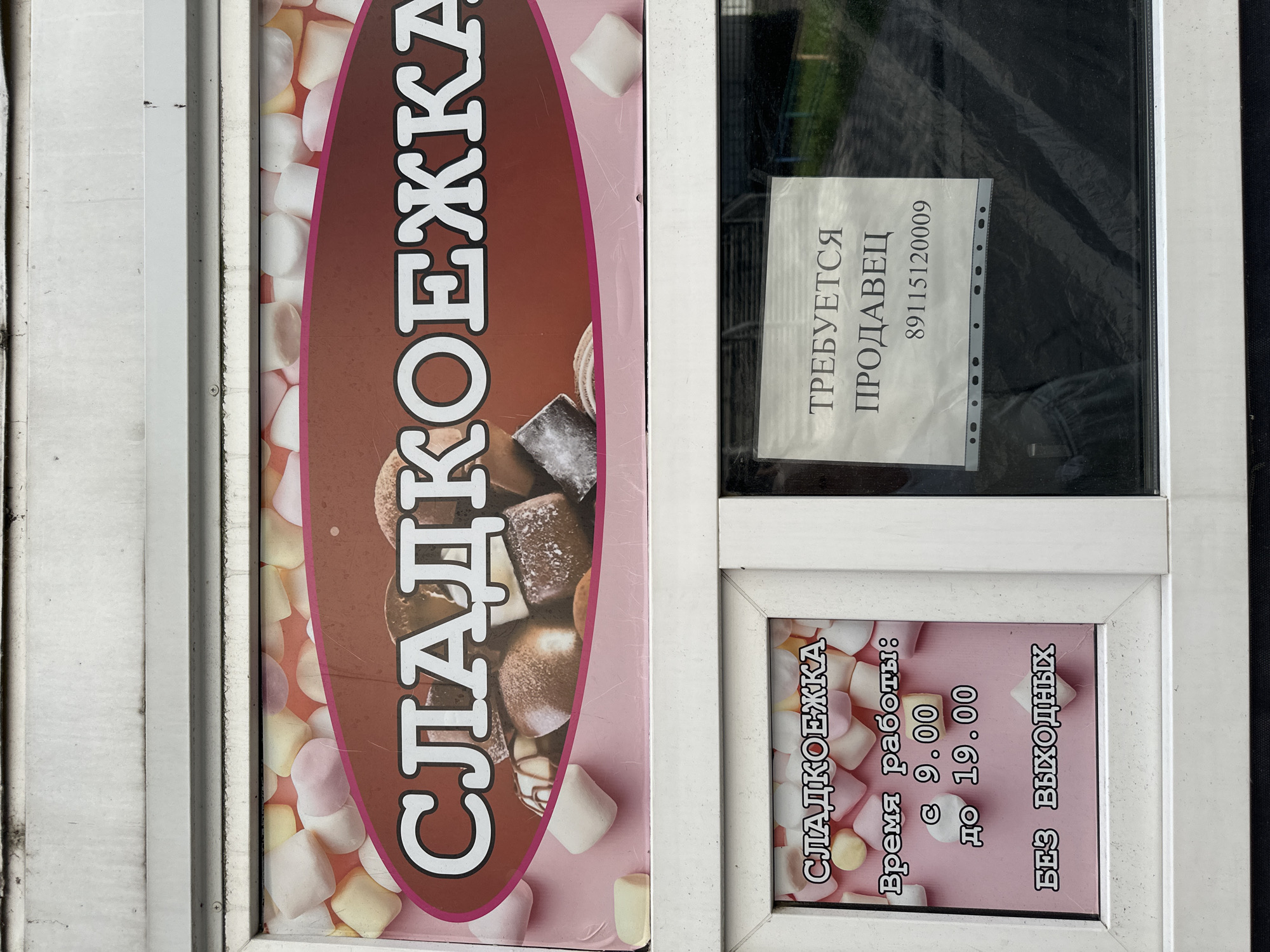 The image shows the storefront of a confectionery shop called "СЛАДКОЕЖКА" (Sladkoezhka). The shop's name is displayed prominently on a large banner with an image of marshmallows. Below the banner, a window displays operational information for the shop. One section reads "Время работы: с 9.00 до 19.00" (Opening hours: from 9.00 to 19.00) and "БЕЗ ВЫХОДНЫХ" (Without days off). Another section of the window displays a sign that says "ТРЕБУЕТСЯ ПРОДАВЕЦ" (Salesperson required) along with a phone number "89115120009". The storefront appears to be located in Rubtsovo, Russia. The overall scene depicts a small retail business with a recruitment notice.