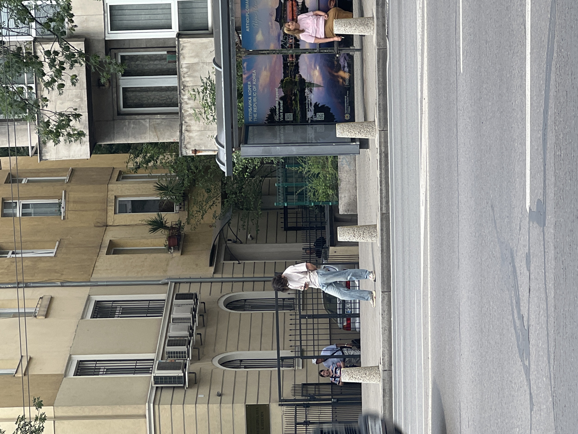The image depicts a street scene in Sofia, Bulgaria, likely near a bus stop. In the foreground, a portion of a street with white lane markings is visible. A young person with dark, curly hair is standing on the sidewalk, looking down at their phone. They are wearing a light-colored t-shirt and light blue jeans. The sidewalk is lined with cylindrical concrete bollards.
Behind the young person, a bus stop shelter is visible, featuring advertisements with images of South Korea. A woman with blonde hair, wearing a pink t-shirt and brown skirt, is seated on the bench inside the bus stop.
Further back, a metal fence separates the sidewalk from a building. Two people are standing near the fence, one appearing to be a woman with a striped shirt, and another person, possibly male, wearing a white cap and a light-colored shirt. A car is parked behind them.
The background consists of multi-story buildings. The building to the left has a light yellow facade with arched windows and multiple air conditioning units installed below the windows. Another building to the right has a more modern concrete facade with balconies. Trees and foliage are present, indicating an urban environment with some greenery. The overall impression is a typical street scene in a European city.