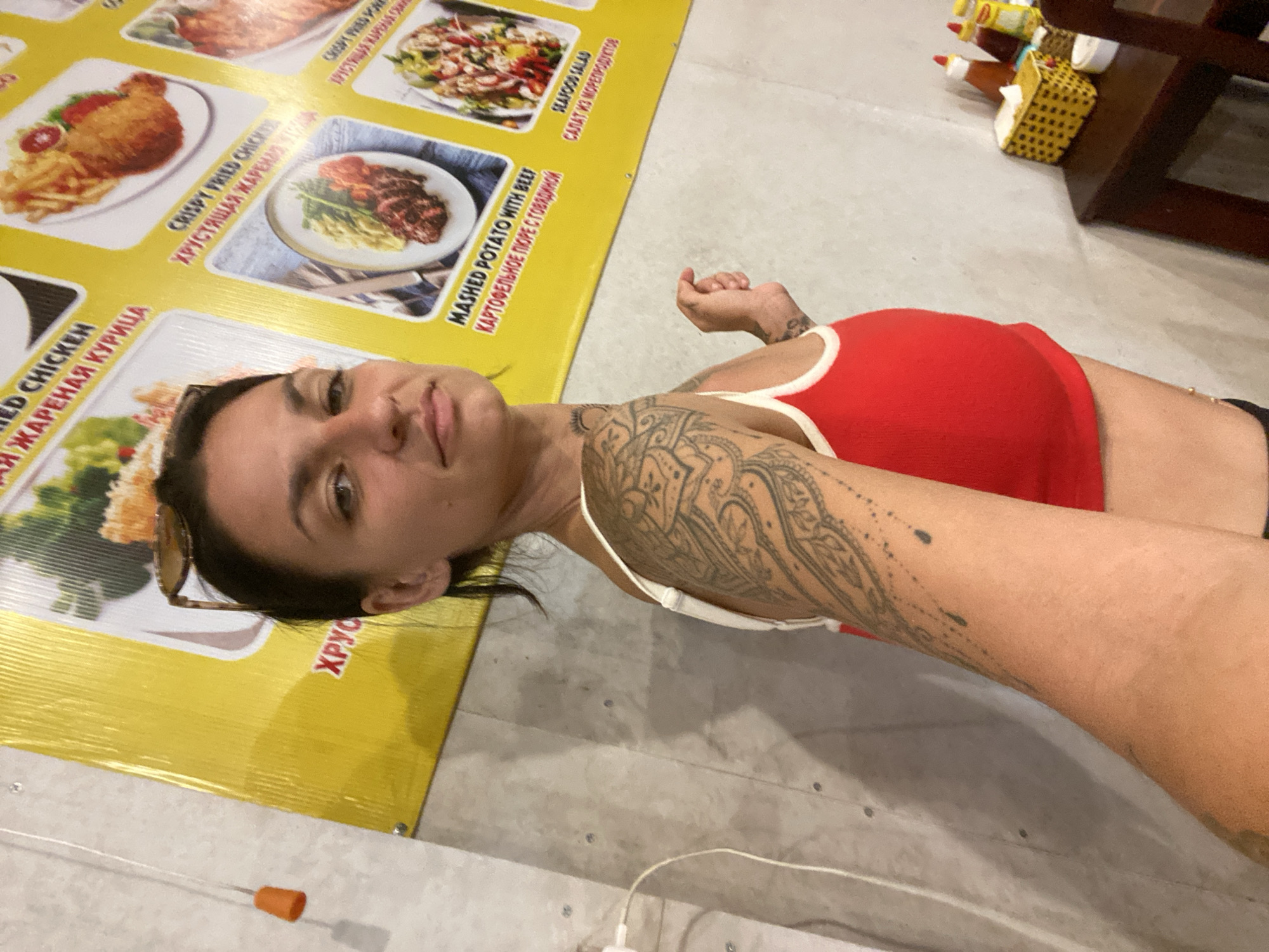 A woman with dark hair, wearing a red tank top and sunglasses perched on her head, stands in front of a yellow menu board displaying food items. Her left arm is covered in a large tattoo. She is looking towards the camera and has her right arm extended, with her hand slightly open. In the background, a wall with visible fixtures and a string with an orange object hanging from it can be seen. A wooden table to the right holds condiments and a tissue dispenser. The menu board displays images of various dishes, including fried chicken and beef with mashed potatoes, and text in English and Cyrillic. The overall scene appears to be an outdoor food stall or restaurant in Phan Thiết, Vietnam.