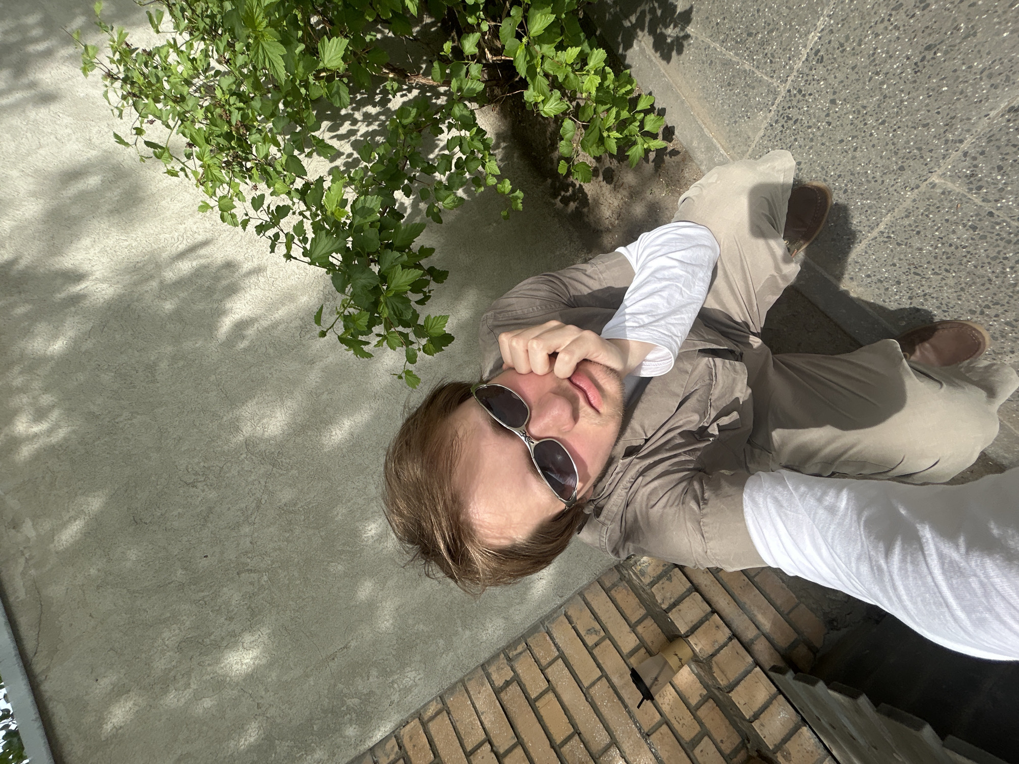 A high-angle shot shows a person wearing sunglasses, a button-up shirt, and khaki pants, lying on a gray concrete surface next to a patch of green plants. The person's right hand is near their face. Part of a brick structure is visible at the bottom of the frame. The location appears to be outdoors in an urban setting.