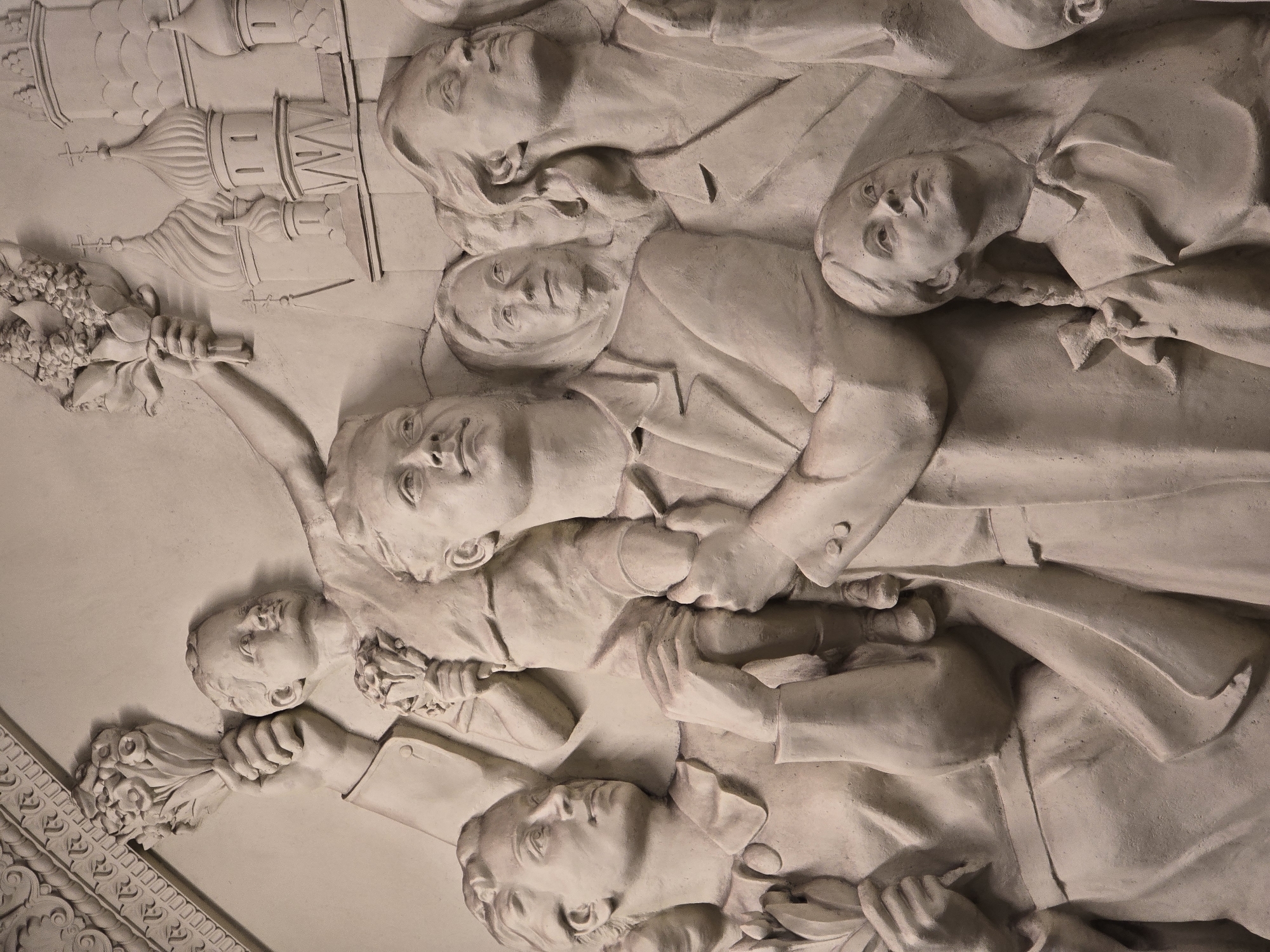 This image shows a bas-relief sculpture. It depicts a group of people, some holding bouquets. A building with multiple domes, reminiscent of Russian Orthodox architecture, is visible on the left side. The sculpture appears to be made of a light-colored stone or plaster. The location is Unknown, Unknown.