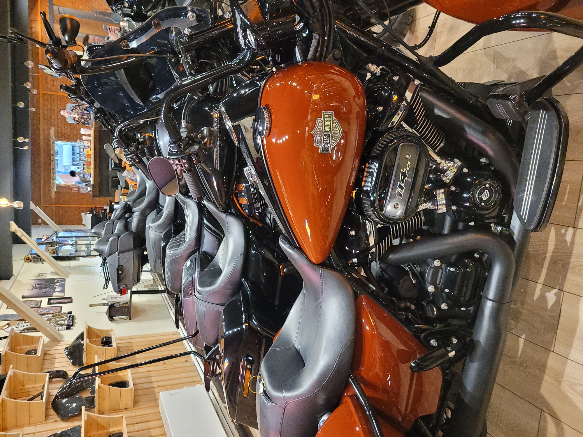 The image captures a display of Harley-Davidson motorcycles in what appears to be a dealership or showroom. Several bikes are lined up, featuring various models with distinctive orange and black color schemes. The motorcycles have detailed engine components visible, including the "114 CI" branded air cleaner cover. The seats are made of black leather, and the fuel tanks bear the Harley-Davidson logo.
In the background, wooden storage bins and racks are visible, holding what seems to be motorcycle accessories or parts. Additionally, there are framed prints and a hanging light fixture, suggesting a stylized retail environment.
While the image provides no explicit indicators of location, it is not possible to confirm the presence of Moscow, Russia.