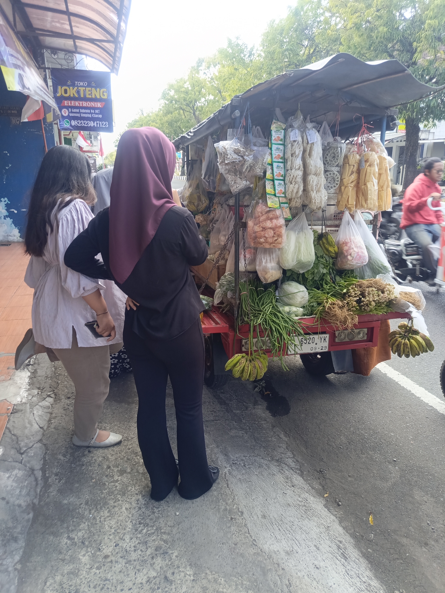 The image depicts a street scene in Desa Gunungsimping, Indonesia, with a focus on a mobile vendor's cart laden with various goods. In the foreground, two individuals are visible. One person, wearing a dark hijab and a black top with dark pants, stands with their back to the camera, right hand on their hip. To their left, another individual in a light-colored, long-sleeved blouse and beige pants is seen from the side, holding a smartphone in their right hand.
The vendor's cart, painted red, is packed with a variety of items, including packaged snacks hanging from the top, bundles of green vegetables like long beans and scallions, cabbages, and bunches of bananas. A license plate on the back of the cart reads "6520 YK 09-29". A store sign above the cart reads "TOKO JOKTENG ELEKTRONIK" with an address and phone number. In the background, a person in a red jacket rides a motorcycle, blurred due to motion. The environment appears to be an urban street with a sidewalk and asphalt road, lined with trees and buildings.