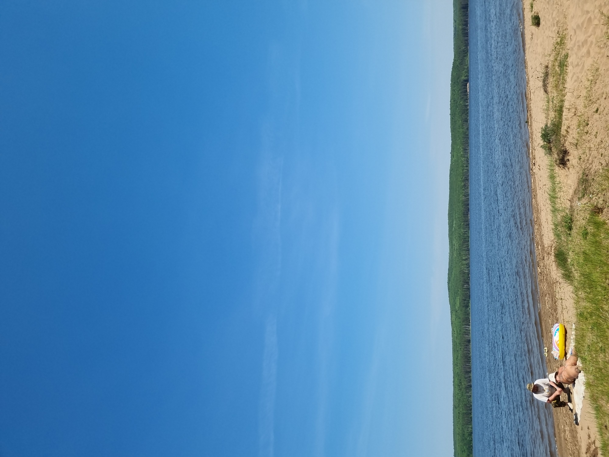 The image shows a bright, clear blue sky occupying the majority of the frame on the left. To the right, there is a shoreline with a body of water alongside a densely wooded area. The shoreline consists of a sandy beach with patches of green grass. On the beach, a person is seated near the water's edge next to an inflatable raft. The location is likely the city of Chaikovsky, Russia.