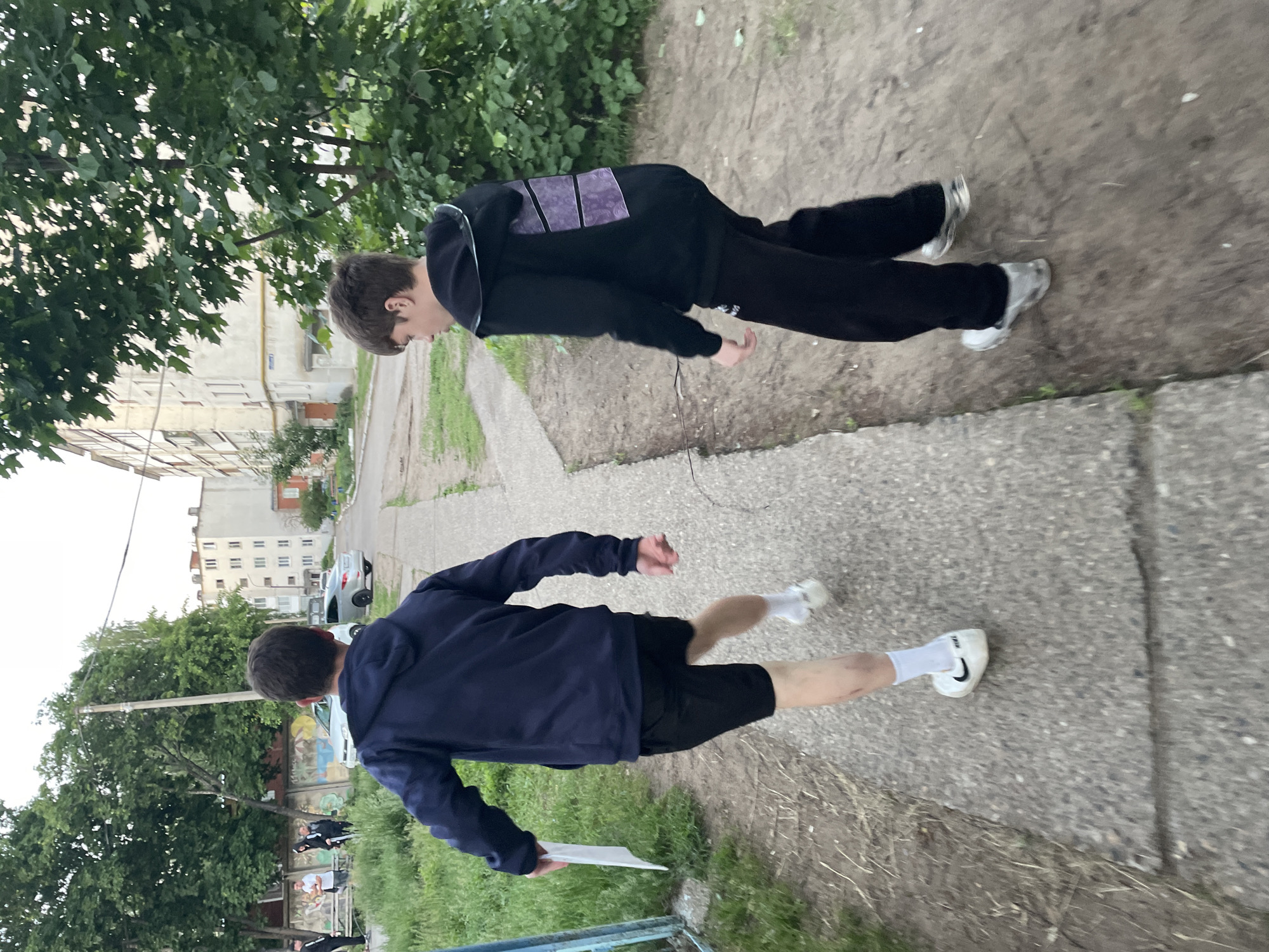 This image captures two young males moving along a concrete path in the city of Noviy, Russia, with an urban residential backdrop. The male in the foreground, seen from behind, wears a dark blue hooded sweatshirt, black athletic shorts, white socks, and white sneakers with a visible Nike logo on the right foot. He holds a white, unfolded sheet of paper in his left hand. His right leg is in mid-stride, displaying a minor abrasion on the shin, suggesting rapid movement or running. The second male, positioned slightly to the right and further back, is viewed from his left profile. He wears a black hooded sweatshirt featuring two rectangular purple patterned panels on the back, black track pants, and light-colored athletic shoes. His head is turned to his left, looking forward. The path they traverse is concrete, showing signs of wear and flanked by a dirt area with sparse vegetation on the right, and green grass and a blue metal gate section on the left. In the background, several light-colored, multi-story apartment buildings, characteristic of Soviet-era architecture, are visible along a street. Two cars, one white and one light grey sedan, are parked on this street. Numerous deciduous trees with green leaves populate the scene, and a utility pole stands between them. In the far left background, a group of approximately four additional individuals stands casually near a low wall or bench. The overall lighting suggests either late afternoon or early evening.