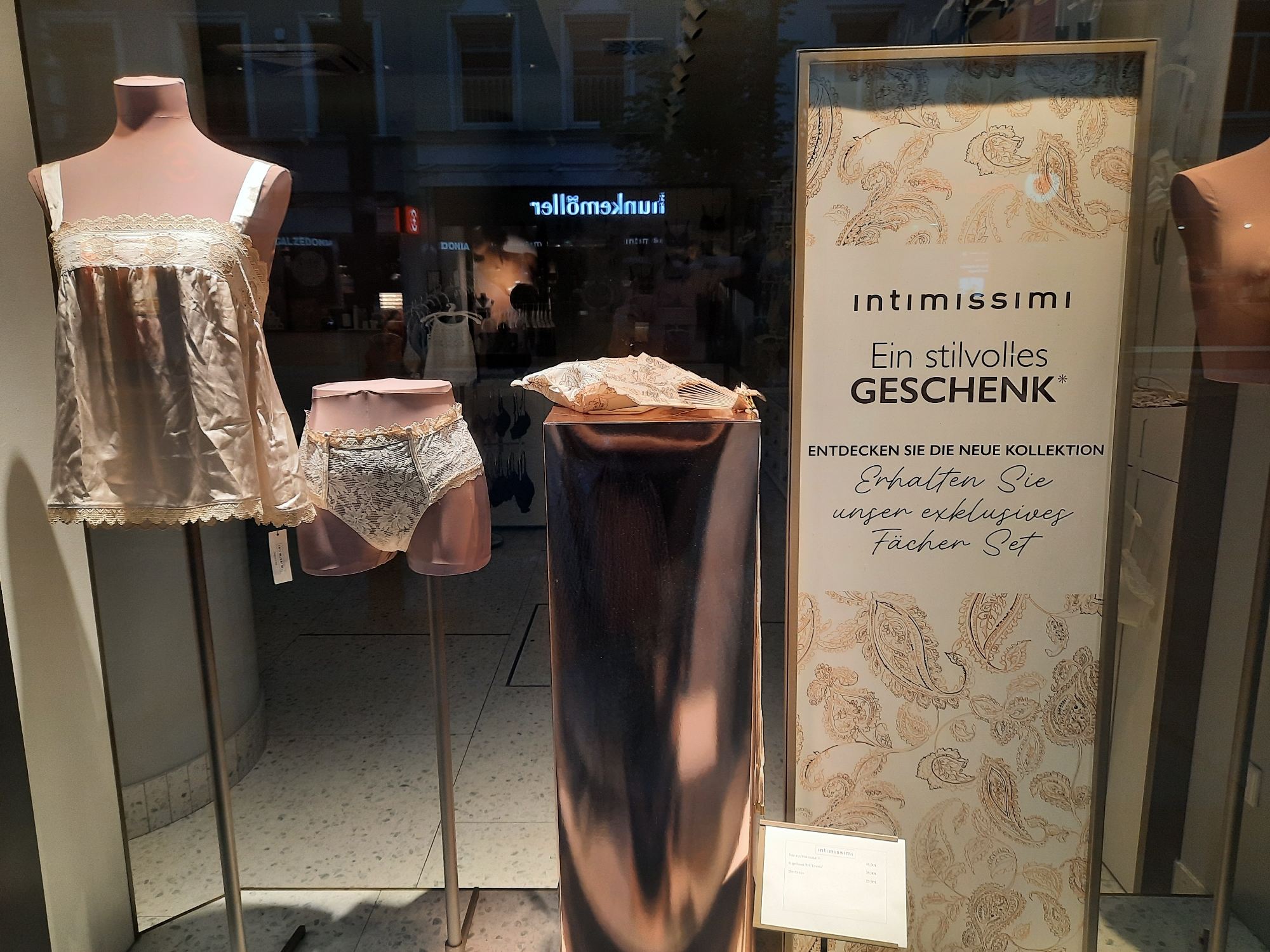The image shows a store window display in Rosenheim, Germany. The display features lingerie on mannequins, with a large sign that reads "Intimissimi" and promotes a stylish gift. The sign also offers a "Fächer Set" (fan set) as an exclusive item. In the background, the reflection of a street with a neon sign can be seen. The overall scene suggests a retail environment, likely a boutique or department store.