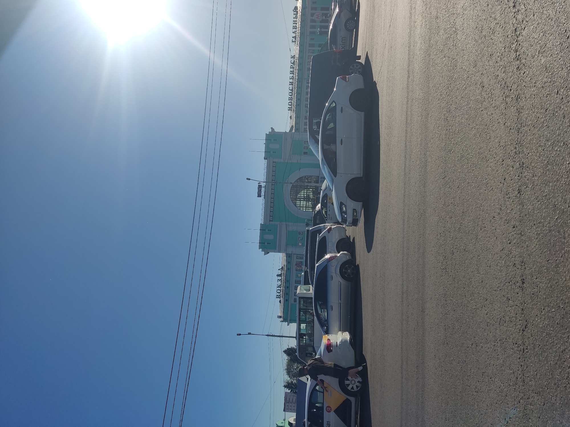 The image captures a daytime scene in front of the Novosibirsk-Glavny railway station in Novosibirsk, Russia. The bright sun is high in a clear blue sky, casting long shadows and creating lens flare. The station building, a prominent turquoise structure with "ВОКЗАЛ" (VOKZAL - Station) and "НОВОСИБИРСК ГЛАВНЫЙ" (NOVOSIBIRSK GLAVNY - Novosibirsk Main) written across its facade, dominates the background.
In the foreground, a traffic jam is evident on the asphalt road. Several cars are stopped, including a white taxi with "581" visible on its side and a yellow logo. A person, dressed in a dark t-shirt and shorts, is standing next to the open driver's side door of the white taxi, possibly interacting with the driver or disembarking. Other vehicles in the congestion include a silver hatchback, a dark-colored van, and other cars whose details are partially obscured. The overall atmosphere suggests a busy urban environment during daylight hours.