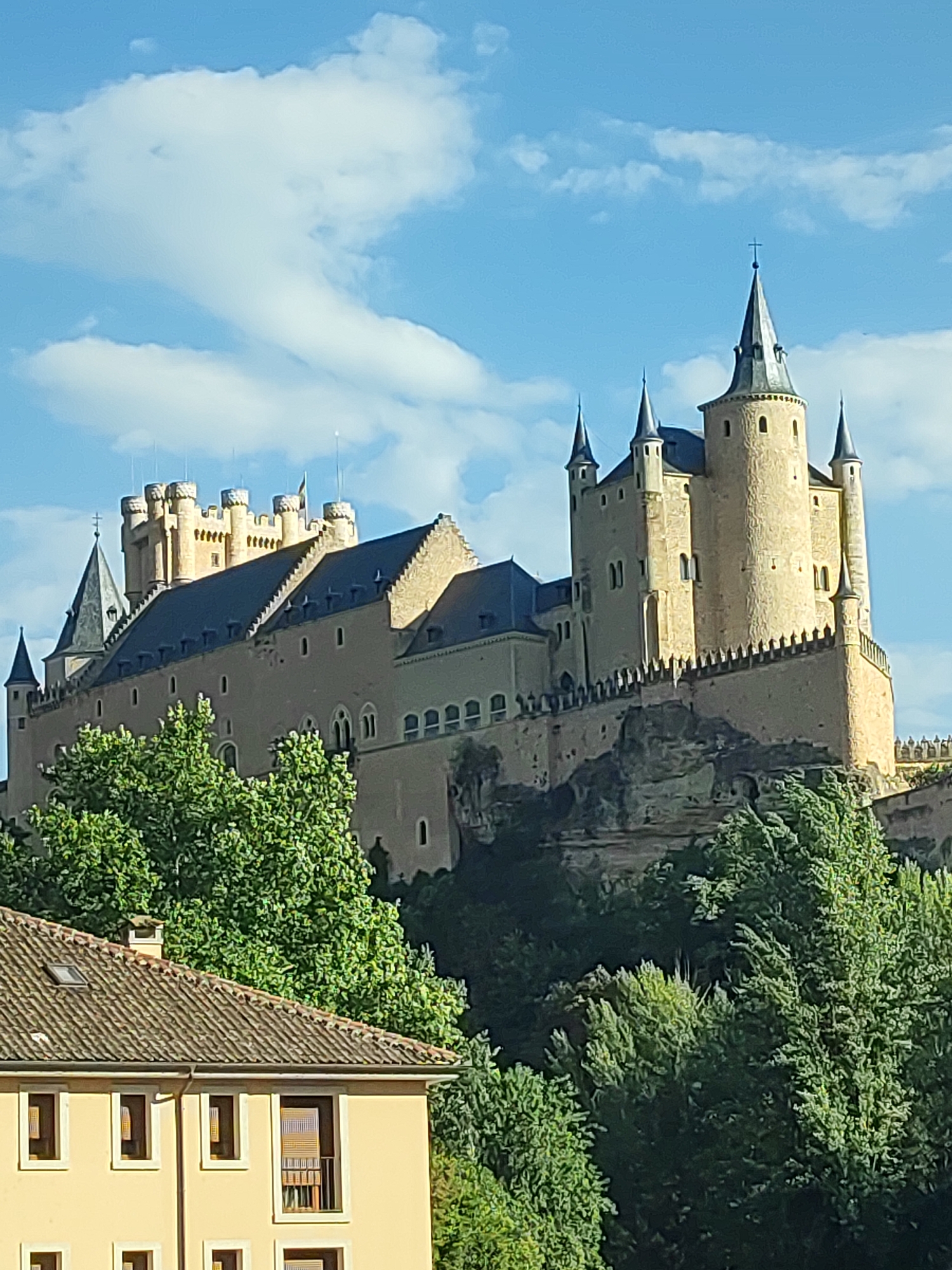 The image shows the Alcázar of Segovia, a medieval castle located in the city of Segovia, Spain. The castle is situated on a rocky promontory overlooking a valley, with lush green trees in the foreground. The sky is a vibrant blue, dotted with fluffy white clouds. The castle itself is a magnificent structure with several towers, turrets, and battlements, constructed from light-colored stone. The main tower, the Torre del Homenaje, is particularly prominent, rising high above the other structures. In the lower left of the image, a portion of a yellow building with a tiled roof is visible, suggesting the presence of other structures in the vicinity of the castle. The lighting suggests it is daytime, with sunlight casting shadows on the castle walls.