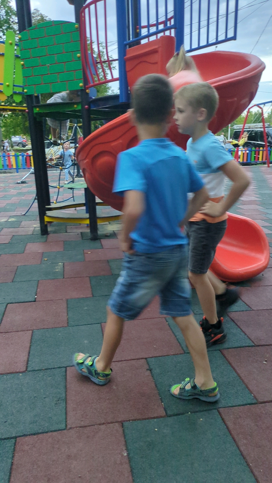 Two young boys are walking on a playground in Chișinău, Moldova. The boy in the foreground is wearing a blue t-shirt and denim shorts, while the boy behind him is wearing a white and orange t-shirt and dark shorts. Both boys are wearing sandals. The playground features a large red slide and other structures made of metal and plastic. The ground is covered with colored rubber tiles. The sky is overcast.