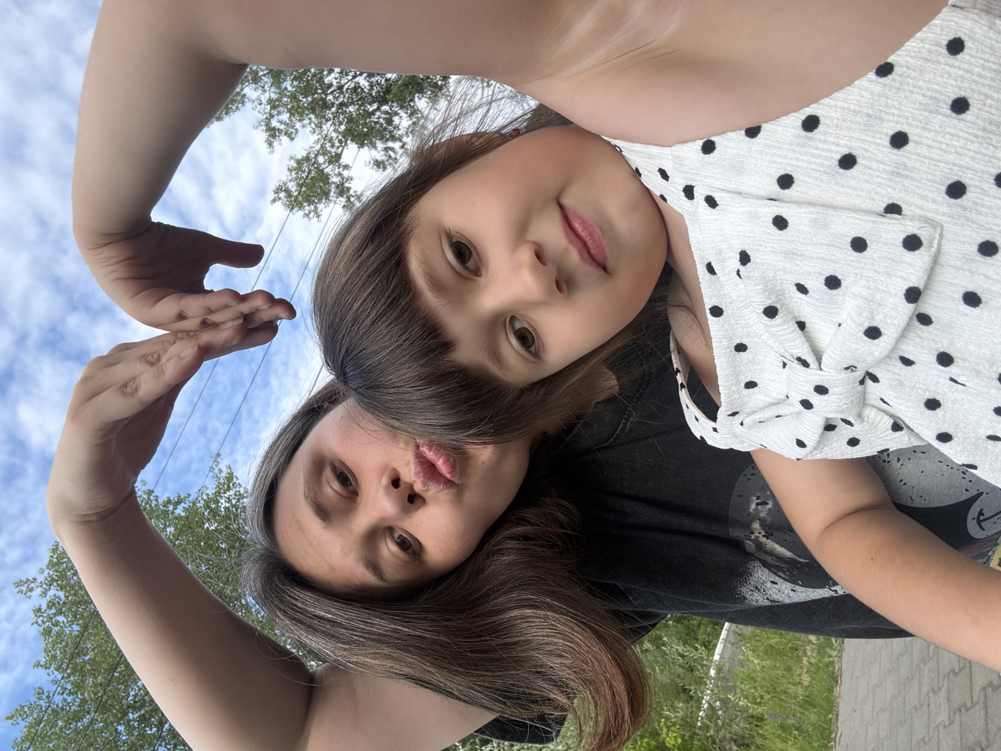 The image is a selfie taken outdoors, likely in Chaikovsky, Russia. Two individuals, an adult and a child, are the main subjects. The adult, in the foreground, is making a kissing face and has their arms raised, with one hand held in front of their face. They have long, brown hair with some gray strands and are wearing a black t-shirt. The child is positioned behind the adult, looking towards the camera with a slight smile. They have dark brown hair and are wearing a white dress with black polka dots. The background shows a blue sky with some clouds, green trees, and a paved surface, suggesting a park or residential area. The lighting indicates it is daytime, likely sunny. The overall mood appears playful and affectionate, given the poses and expressions of the subjects. There is no visible text in the image.