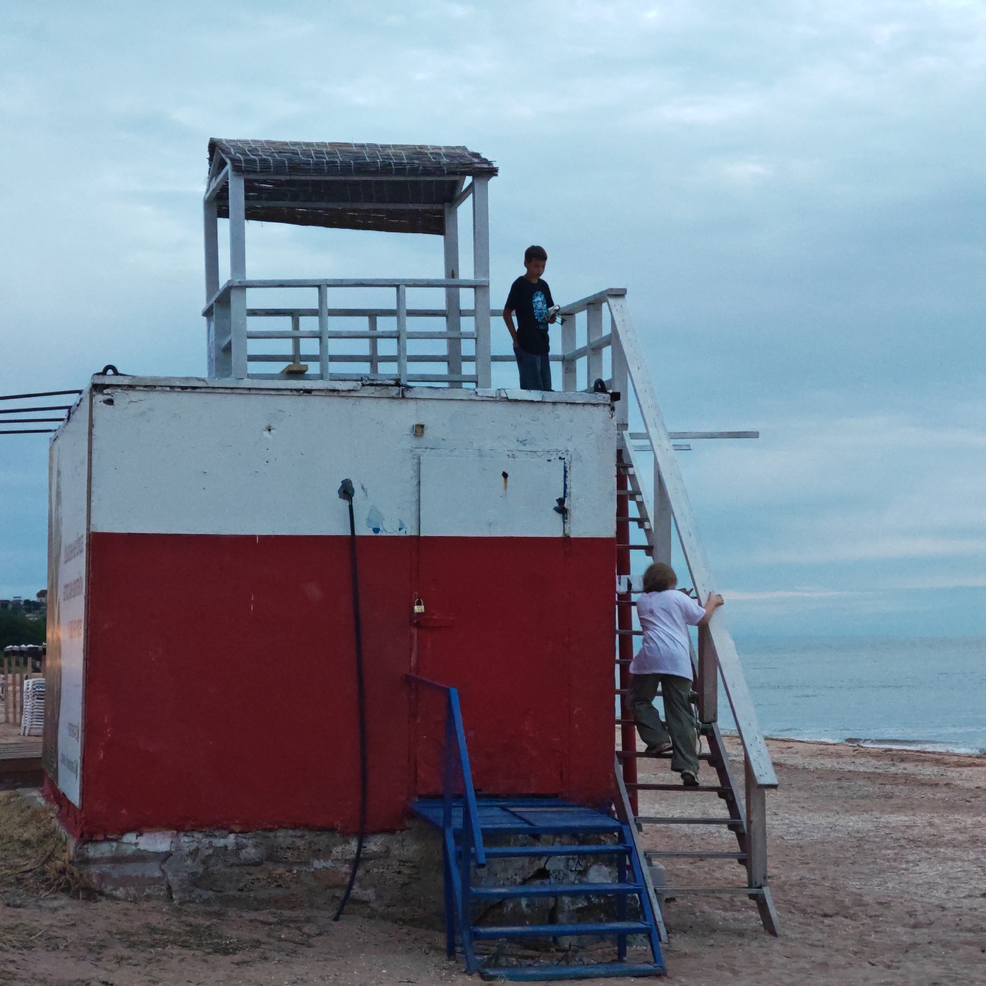 The image depicts a two-story lifeguard or observation tower situated on a sandy beach under an overcast, pale blue-grey sky. The lower section of the tower is painted red, while the upper section is white, topped with a slatted wooden roof.
Two individuals are present around the tower. A boy, wearing a dark t-shirt and jeans, stands on the upper platform, looking downwards. Another person, seen from behind, dressed in a light-colored t-shirt and dark pants, is ascending the exterior wooden staircase that leads to the upper platform. A separate blue metal staircase provides access to the lower red section of the building.
The setting is a beach with light brown sand, sparsely covered with shells, extending towards the calm ocean visible to the right. Small ripples or waves are noticeable near the water's edge. The soft, diffused light from the cloudy sky suggests either early morning or late afternoon.
Visible details on the tower include a black cable running vertically down the white section, a padlock on a red door on the lower level, and a rectangular white sign on the left side of the red section, though its text is unreadable. The base of the structure reveals concrete and brickwork. The scene is located in a coastal area, specifically in городское поселение Щелкино, Ukraine. No specific emotions are discernible from the individuals.