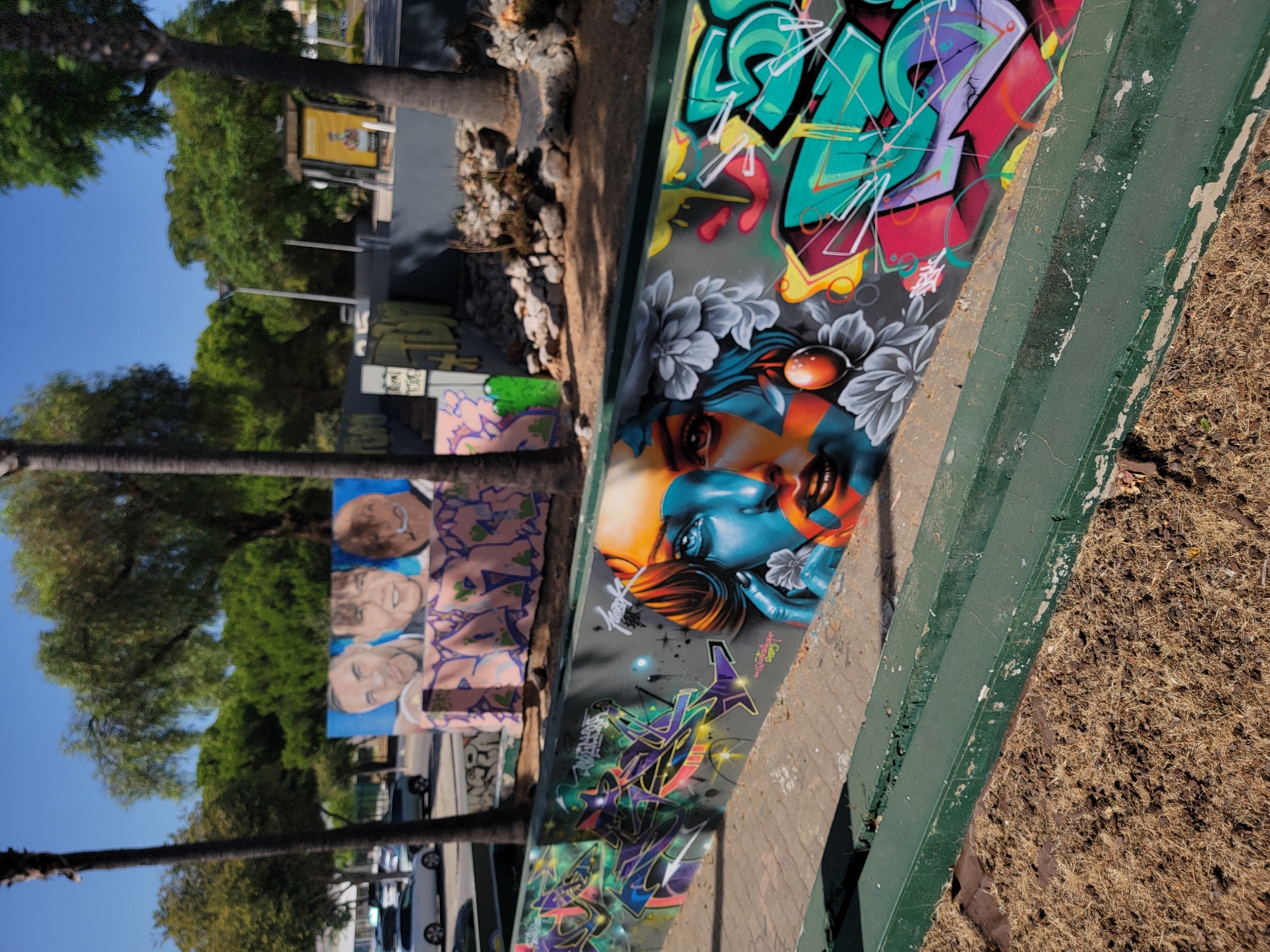 The image depicts a section of urban street art in Quarteira, Portugal. In the foreground, a low, weathered green barrier separates a patch of dry, brownish ground cover from a paved walkway. The walkway is adorned with vibrant graffiti. Dominating the center of the graffiti is a striking mural of a woman's face, rendered in blues, oranges, and reds, with a single red balloon-like object near her mouth and grey floral motifs surrounding her. To the left of the woman's portrait, abstract graffiti in shades of purple, green, and yellow is spray-painted onto a dark surface. To the right, more colorful graffiti, primarily in teal, pink, and yellow, features bold, stylized lettering. In the background, behind a wall also covered in graffiti, are palm trees, dense green foliage, and a clear blue sky. A few cars are visible on a street to the left, and what appears to be a public information kiosk stands further back on the right. The scene suggests an outdoor public space, possibly a park or a promenade, decorated with contemporary street art.