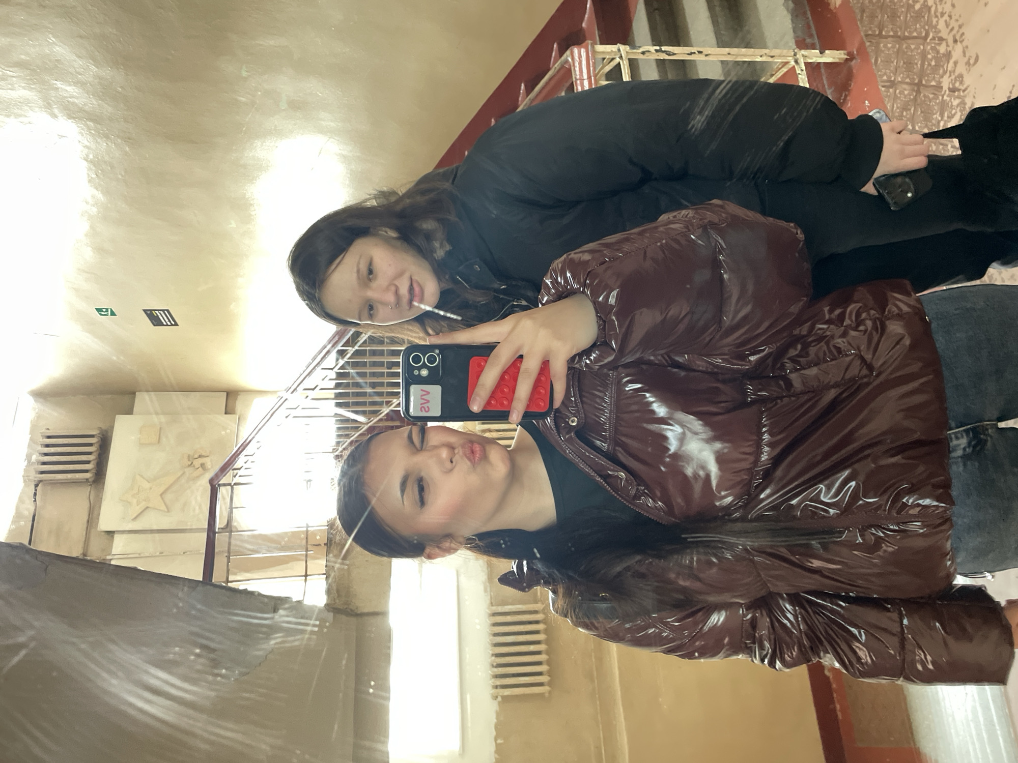 Two young women are seen in a mirror selfie, positioned in what appears to be an entranceway or corridor of a building. The woman in the foreground wears a shiny, brown puffer jacket and is holding a black smartphone with a red case. She has dark hair and is making a pouting expression. The second woman stands behind her, wearing a black puffer jacket, and she has a stick or small object in her mouth. The background shows a staircase with a red railing, beige walls, and architectural details including a star-shaped design on the wall. The setting is characteristic of the interior of a building in Saint Petersburg, Russia, considering the architectural style and general appearance.