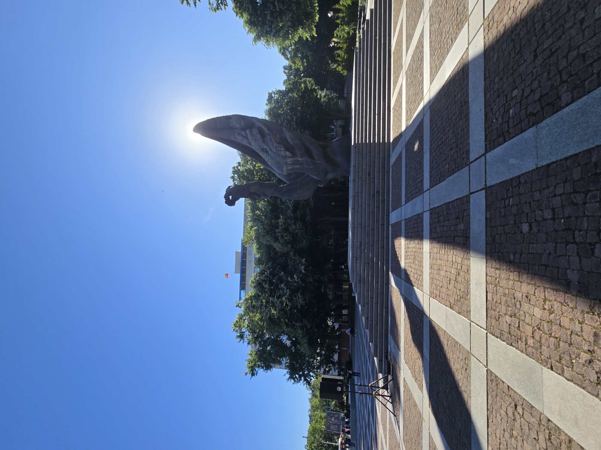 The image shows a large, dark-colored statue of an eagle with its wings spread against a bright blue sky. The sun is positioned directly behind the eagle, creating a silhouette effect. Below the statue is a landscaped area with trees and a building in the background. The ground is paved with a geometric pattern of light and dark stones. In the lower part of the image, there is a temporary structure, possibly for an event, with speakers and signage. The image is taken at the monument to the Silesian Insurgents in Katowice, Poland.