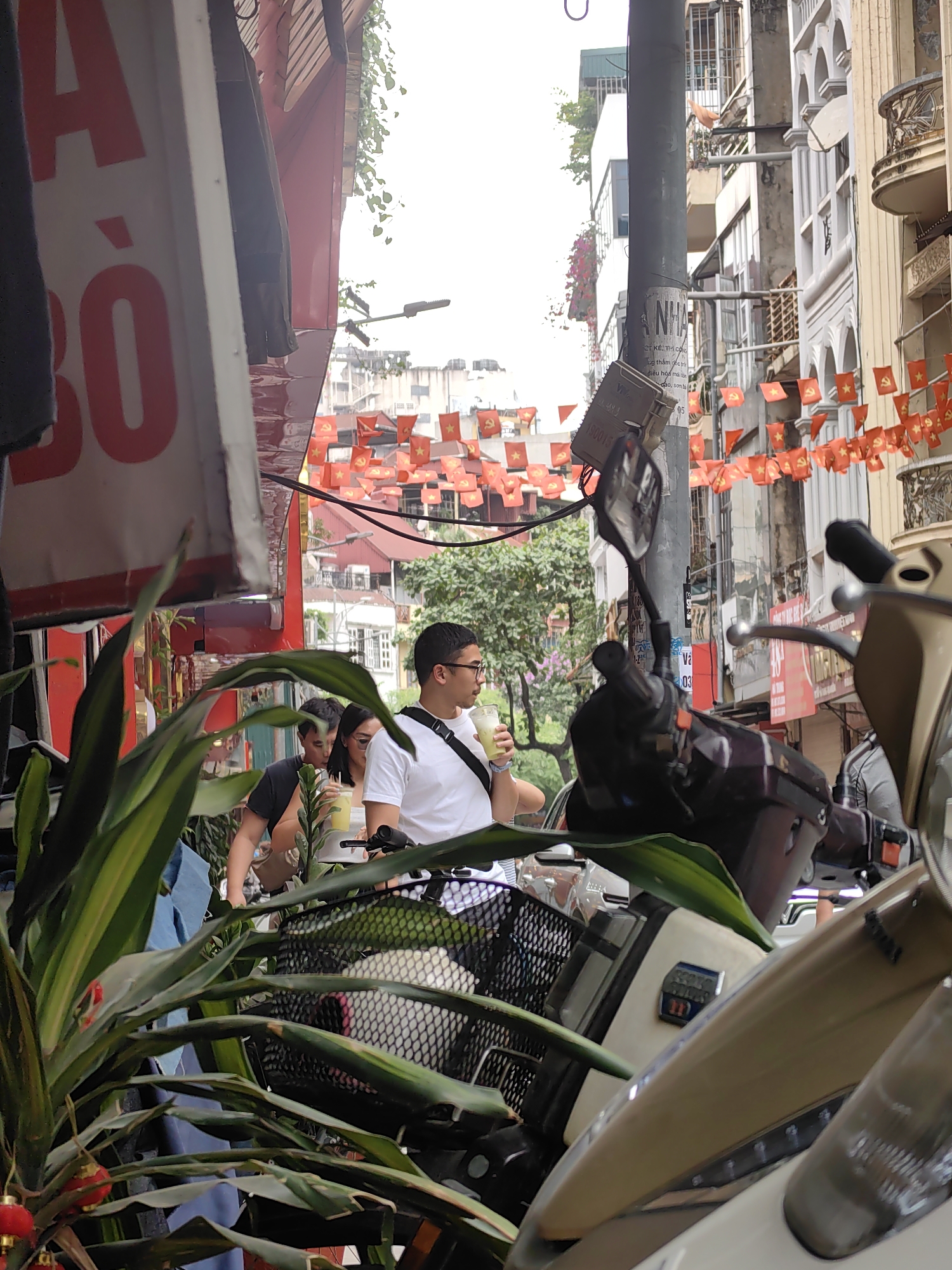 This image depicts a vibrant, busy street scene in Hà Nội, Vietnam, under a bright, likely overcast, daytime sky.
In the immediate foreground, large green leaves of a plant partially obscure the view, featuring small red decorative ornaments or lanterns. Parts of a scooter or motorcycle, including a basket, handlebars, and beige bodywork, are also visible. To the left, a large red and white sign prominently displays the Vietnamese text "A BÒ", possibly indicating a "Phở Bò" (beef noodle soup) establishment or a shop selling beef.
The midground features several people and additional vehicles. A man wearing glasses, a white t-shirt, and a black shoulder bag stands with a clear drink in hand. Behind him, other individuals, including a person in a black t-shirt and a woman wearing sunglasses, are partially visible, suggesting a casual street environment.
The background reveals a dense urban setting with multi-story buildings lining a narrow street. Overhead, numerous small, rectangular red flags with yellow stars, characteristic of Vietnam's national flag or the Communist Party flag, are strung across the street, adding a distinct cultural and political element to the scene. Street poles with various stickers displaying partial Vietnamese text like "ANH NHÀ", "CT KẺ, THÌ CÓ", "ng thấm gốc t", "điều hòa mái tê", and numbers such as "190015", are also present. The overall impression is one of a bustling daily life in a Vietnamese city.