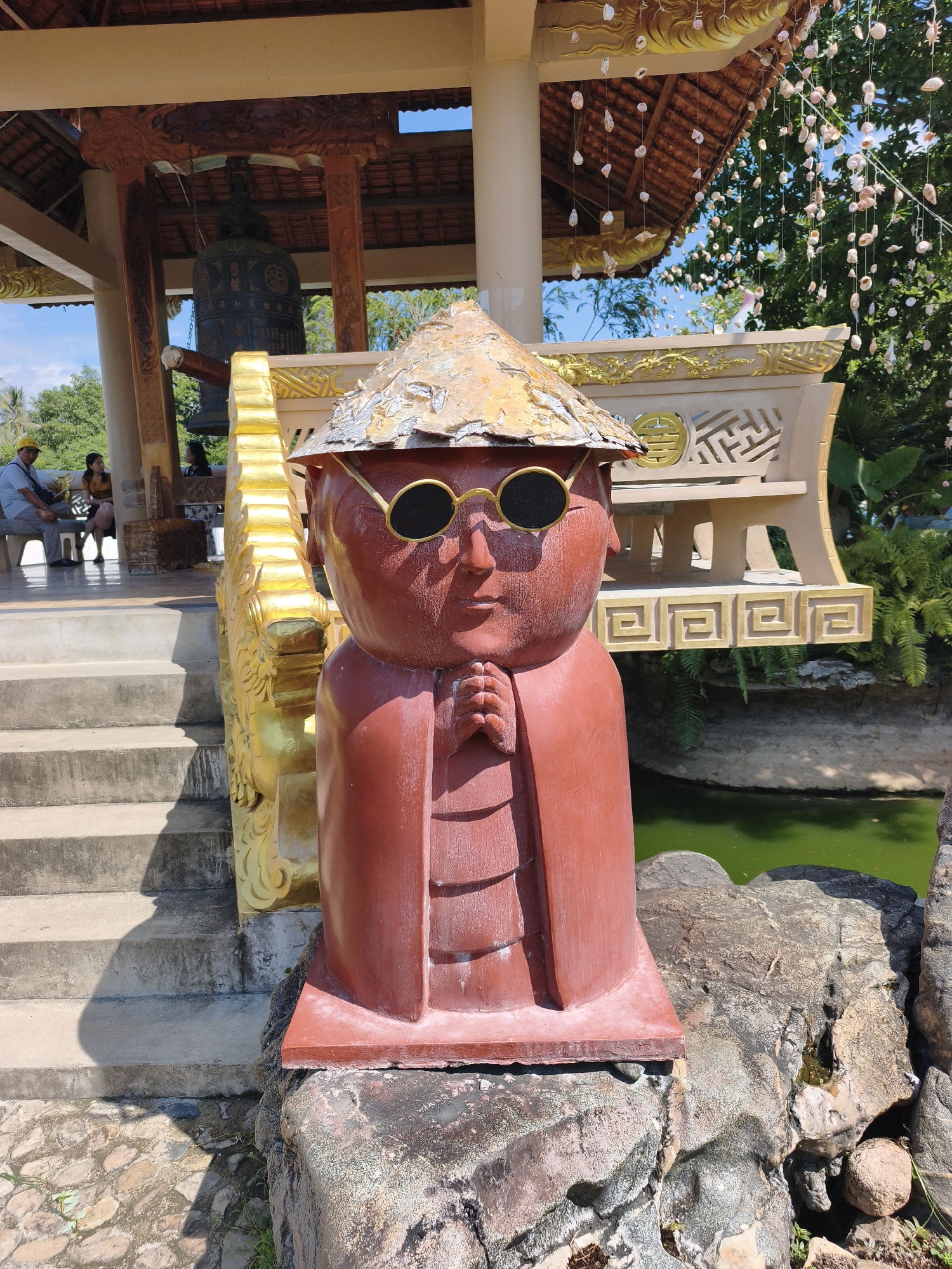 A close-up, eye-level shot shows a reddish-brown statue with a conical hat and round sunglasses in the foreground. The statue is positioned in front of a body of green water and is adorned with gold detailing on its hat and robe. Behind the statue, a large bell is visible under a tiled roof supported by a wooden structure. In the background, a few people are seated on a bench, and lush green trees and blue sky fill the upper portion of the frame. The scene appears to be located in a temple or garden setting.