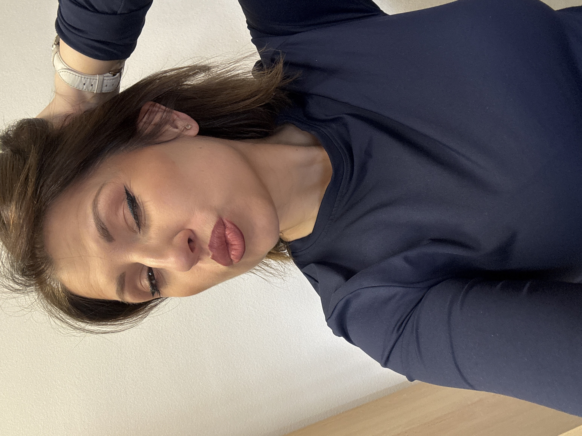A woman is posing for a selfie. She is wearing a long-sleeved, dark blue shirt. She has brown hair, fair skin, and is wearing makeup, including eyeliner and lipstick. A white watch is visible on her left wrist. The background consists of a white wall and a light brown surface. The location is indeterminate as it is not possible to infer the city or country from the visual information.