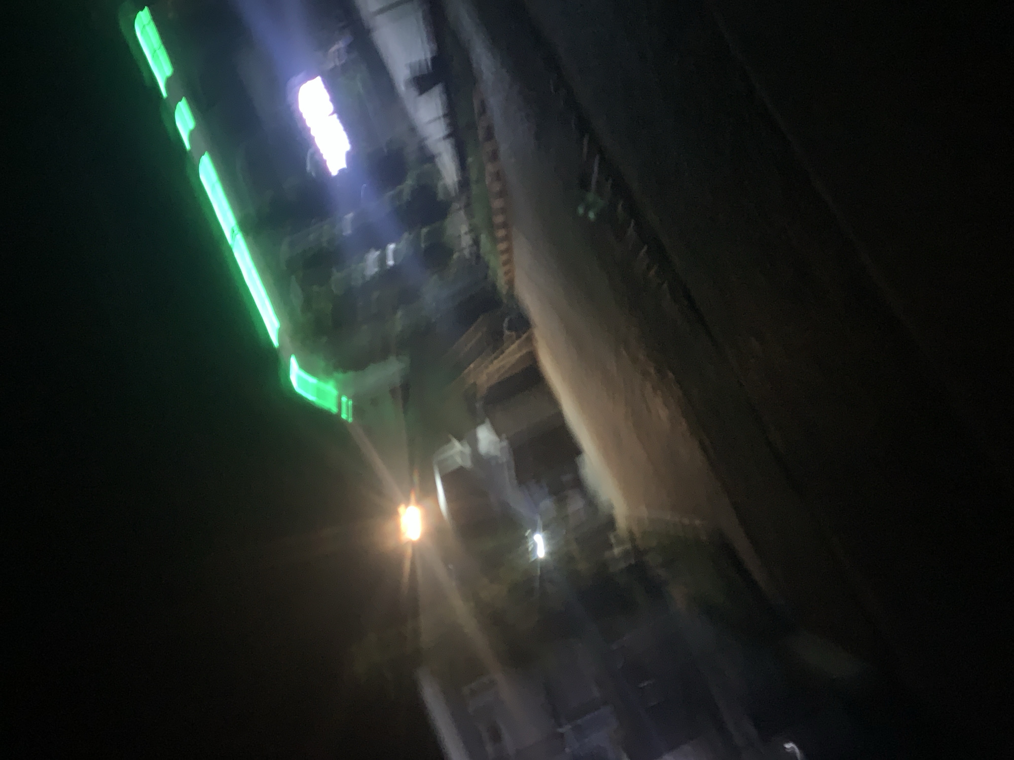 The image is a blurry, low-light photograph taken at night, likely from a moving vehicle, showing a street scene in Jalingo, Nigeria. A prominent feature is a bright green neon sign stretching diagonally across the upper portion of the frame. Below the neon sign, a lighter, rectangular sign with white or light purple lettering is visible. In the foreground and midground, there are indistinct shapes that appear to be buildings and a paved road. A streetlamp with a bright orange glow is visible in the left midground. The overall impression is one of a commercial or entertainment district at night, with blurred motion contributing to the abstract quality of the image.