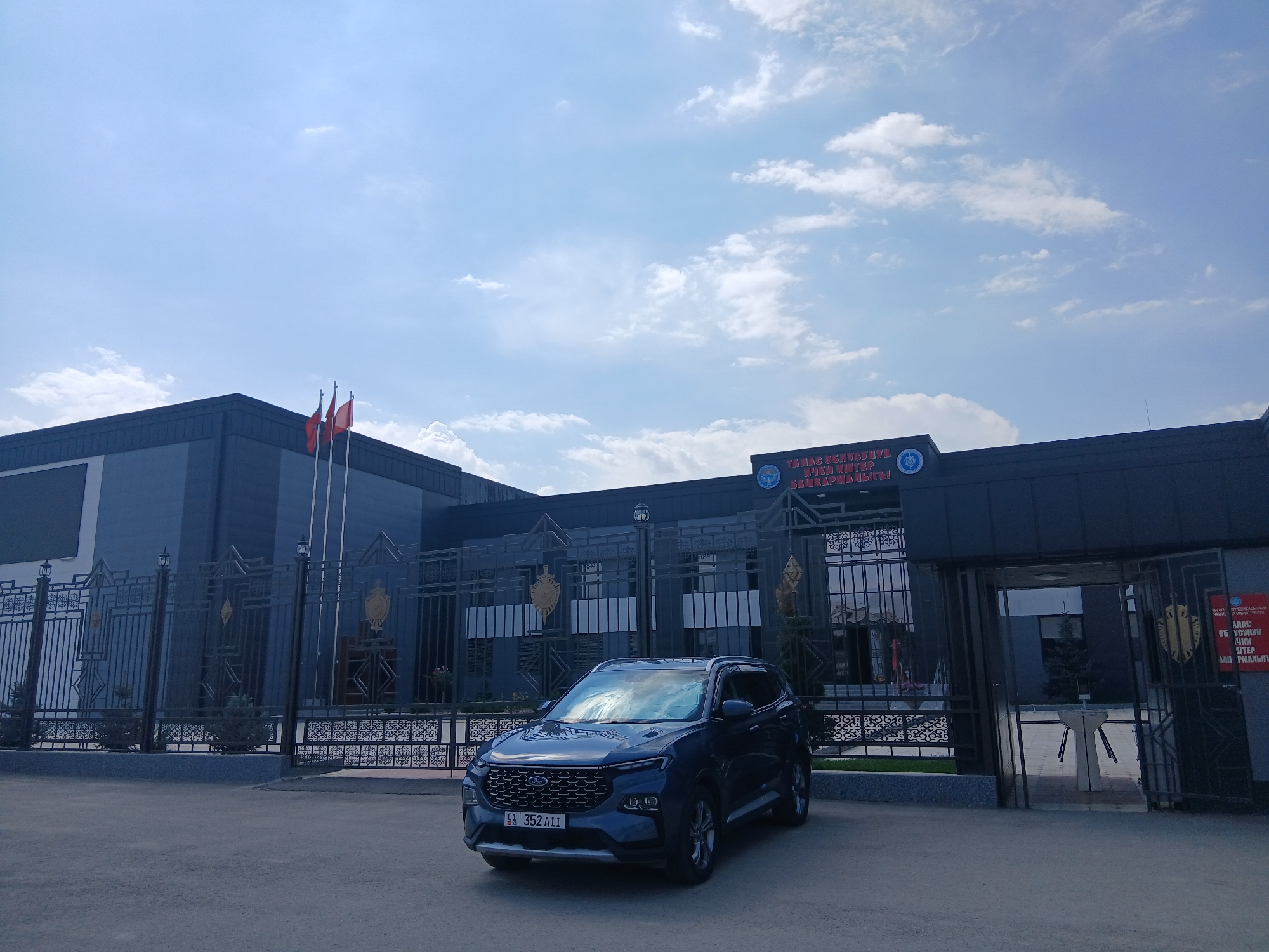 In front of a dark-colored modern building with a large ornamental metal fence, a dark blue Ford SUV is parked on an asphalt surface. The building has the text "ТАЛАС ОБЛУСУНУН" and "БАШКАРМАЛЫГЫ" visible in red lettering above its entrance. Three red flags are mounted on poles to the left of the building's entrance. The sky is blue with scattered white clouds. The image appears to have been taken during the daytime.