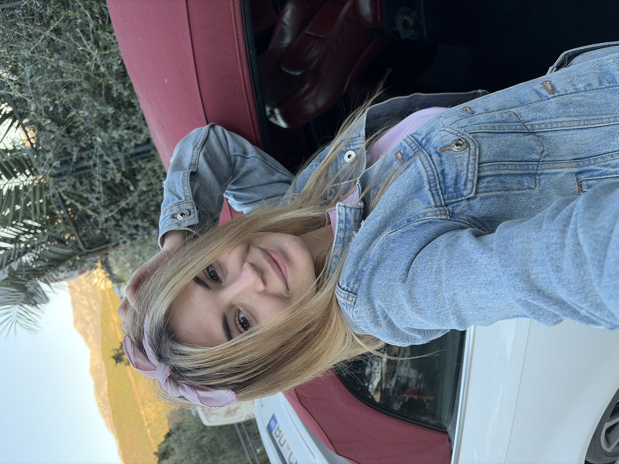 A woman poses in the front passenger seat of a convertible car. She wears a light blue denim jacket over a pink top and a pink ribbon in her hair. The car has red leather seats, and the exterior is white with a red top. Outside the car, there is greenery. In the background, a mountain and sky can be seen. A partially visible license plate is seen on the car, which reads "BU." The location is consistent with the scenery around Budva, Montenegro.