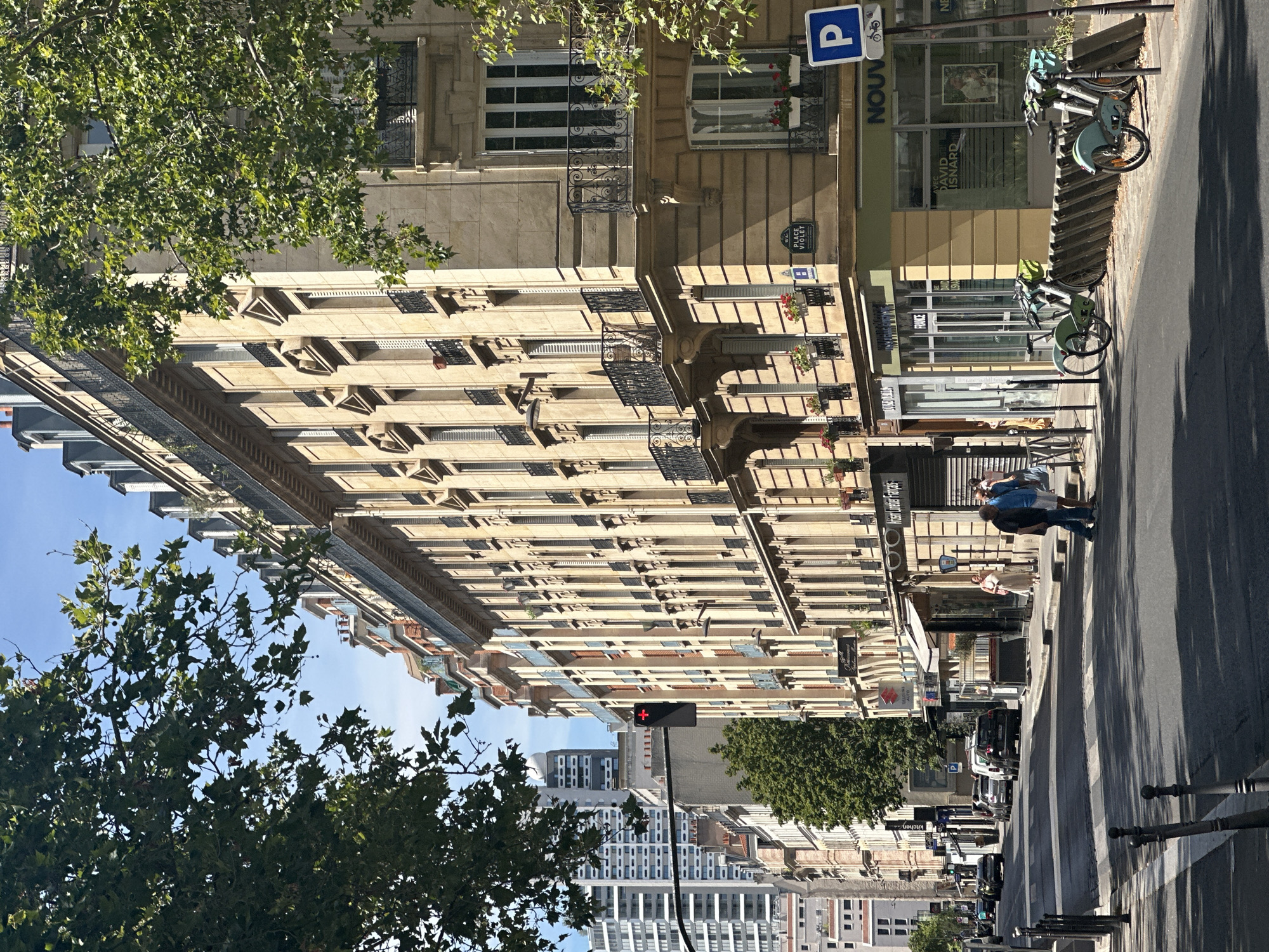 The image captures a street scene in Paris, France, under a clear blue sky. The foreground shows a sun-drenched asphalt street with a pedestrian crossing marked by white stripes. Several dark metal posts line the side of the road. On the right side of the street, a row of pale yellow apartment buildings with ornate architectural details stands tall. These buildings feature multiple stories with regularly spaced windows, many equipped with dark metal balconies and shutters. Some windows have flower boxes with red flowers. A blue and white parking sign with a bicycle symbol is visible on a pole. Next to it, a weathered wooden barrier supports two bicycle racks, with one light blue bicycle and one green bicycle docked. Shops are visible at the ground level of the buildings, with signage including "PLACE VIOLET" and "NOUV."
On the sidewalk in front of the buildings, a few people are present. Two men in casual attire are standing on the sidewalk, one in a dark blue shirt and the other in a lighter blue shirt and shorts. Further down, a woman in a light-colored dress is walking. Across the street, several cars are parked and driving, including black SUVs and a silver sedan. A traffic light with a red signal is suspended over the intersection. In the background, taller modern buildings with white and gray facades rise above the Parisian architecture, suggesting an urban environment. Lush green trees with dappled sunlight filtering through their leaves partially frame the scene from the top left and right.