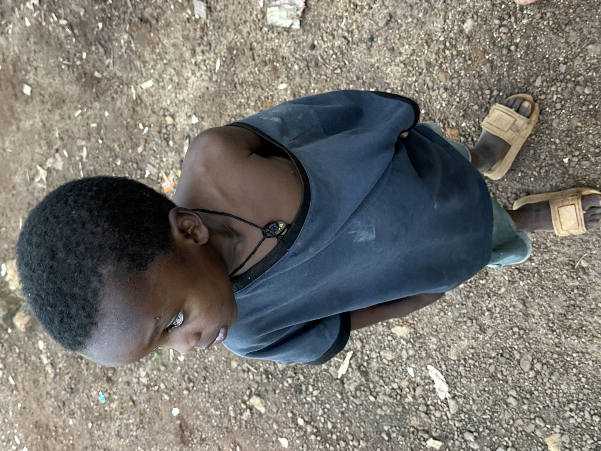 A high-angle medium shot shows a young child standing outdoors on a dirt ground in Gura Top, Nigeria. The child, who appears to be a boy, is looking to his left with a pensive expression. He has short, dark hair and his head is tilted slightly. He is wearing a dark blue t-shirt that is off one shoulder, revealing his collarbone and upper chest. Around his neck is a dark cord with a pendant. His feet are bare, except for a pair of light brown slides on his right foot. The ground beneath him is composed of small stones and dirt, with some scattered debris including bits of plastic and paper. The background is blurred, suggesting a natural or undeveloped environment.