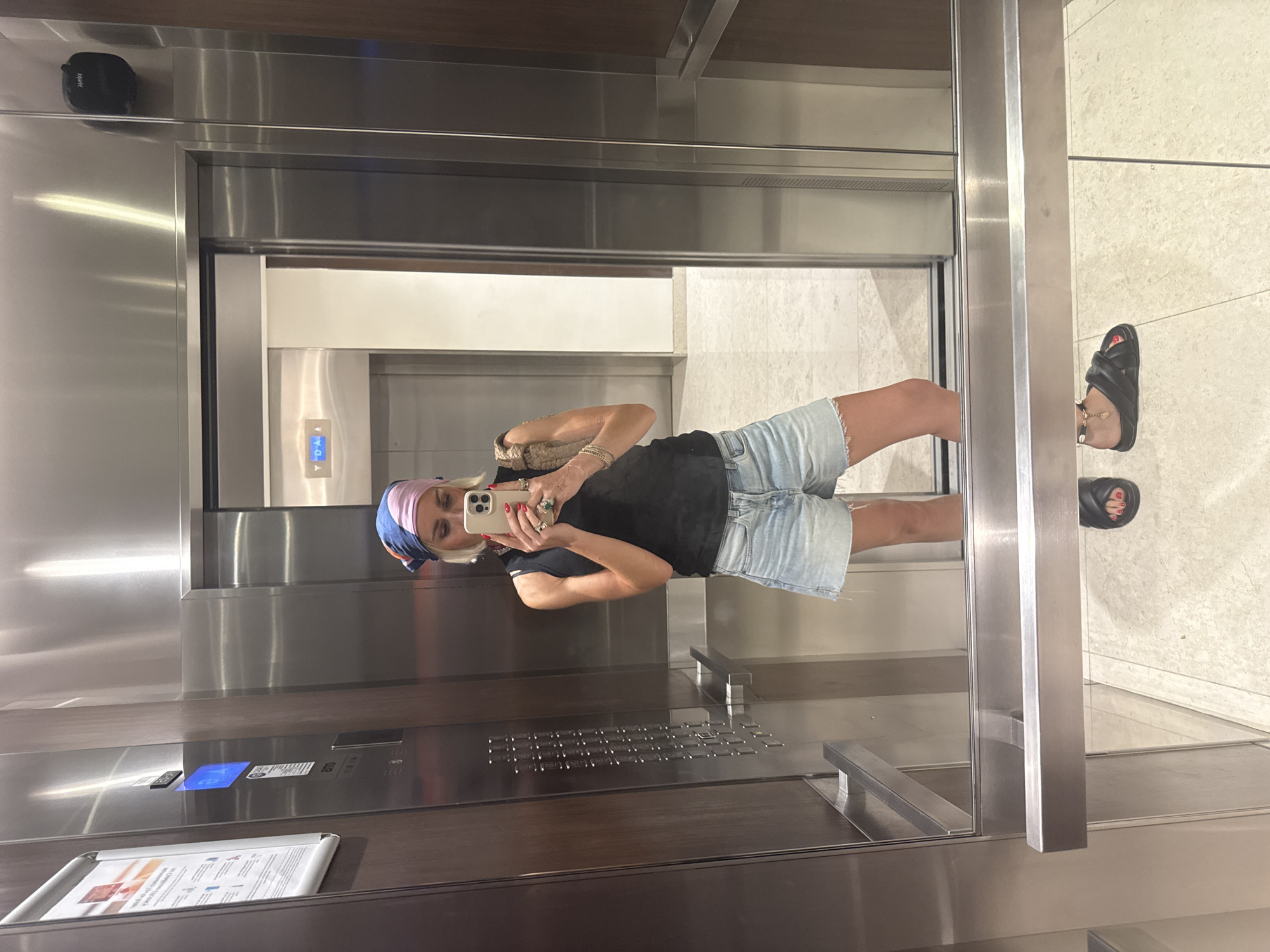 The image shows a woman taking a mirror selfie in an elevator. She is wearing a black tank top, denim shorts, a colorful headscarf, and black slides. She is holding a phone in her right hand and has several rings on her fingers. The elevator has stainless steel walls and a wooden accent panel. The floor is tiled with a light-colored marble pattern. The elevator doors are open, revealing a glimpse of the hallway beyond. The scene is likely in a modern building in Dubai, United Arab Emirates, given the typical aesthetics of such locations.