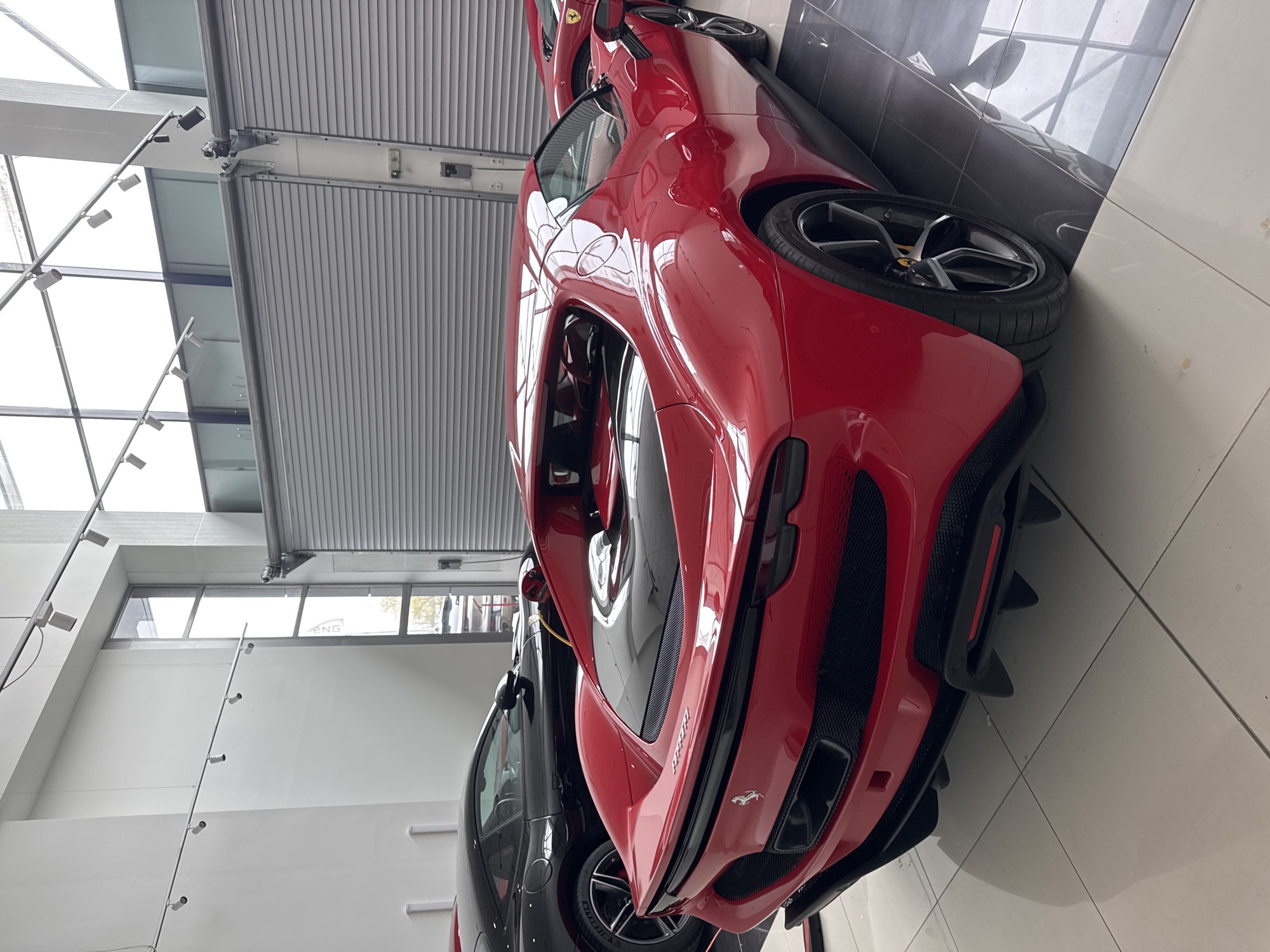 The image shows the rear of a red Ferrari sports car inside a showroom. Adjacent to it is a black car, and other vehicles are visible in the background. The showroom has a white tiled floor, and the walls are white with large windows and a closed garage door. The architecture includes exposed structural supports with lighting fixtures. The location might be in Sofia, Bulgaria.