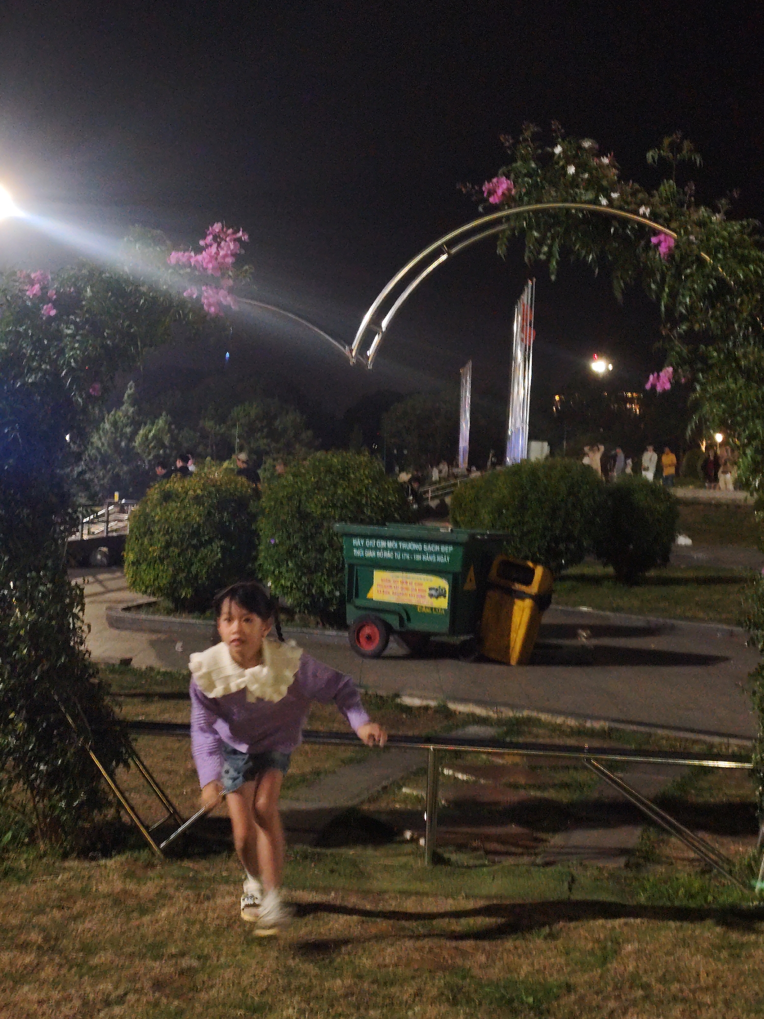 The image captures a nighttime scene in Da Lat, Vietnam, likely at a park or recreational area, given the presence of manicured bushes, lampposts, and decorative floral arches. In the foreground, a young girl with dark hair styled in pigtails is captured mid-stride, appearing to be running or playing. She is wearing a purple long-sleeved shirt with a large, ruffled white collar and denim shorts, paired with white sneakers. Behind her, a green trash receptacle on wheels, with Vietnamese text visible, stands next to a yellow trash can. In the mid-ground, several groups of people are visible, some standing and others walking, illuminated by artificial lighting. The background reveals more foliage, flags or banners on tall poles, and what appears to be a light fixture emitting a bright beam of light. The overall ambiance is one of outdoor evening activity.