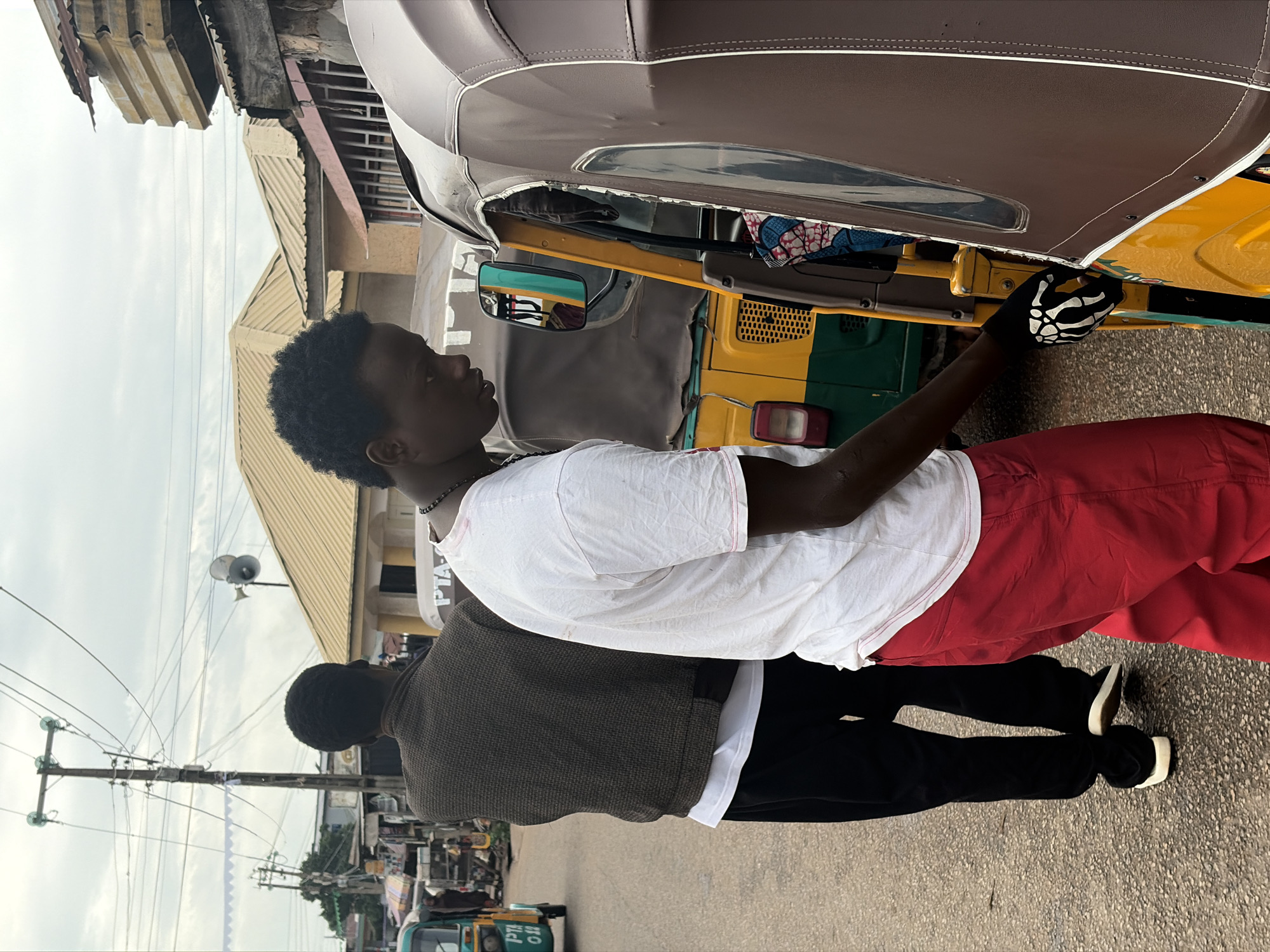 In Jos, Nigeria, two men are standing on a paved street. The man in the foreground is wearing a white t-shirt and red pants, with a black glove on his right hand. He is leaning towards a brown and yellow auto-rickshaw, with his hand on the vehicle. Behind him, another man is wearing a patterned brown jacket over a white t-shirt and black pants. He is walking away from the camera. In the background, several auto-rickshaws are parked along the street, and buildings with corrugated metal roofs are visible. Power lines and poles run overhead. The sky is overcast.