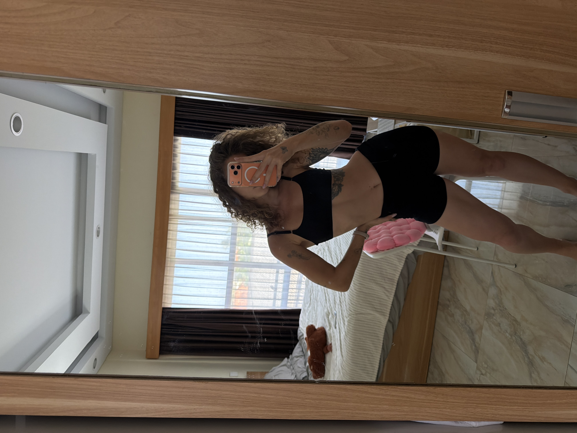 A woman with curly brown hair is taking a selfie in a full-length mirror. She is wearing a black sports bra and black shorts. She has tattoos on her arms and stomach. In the reflection, a bed with white and striped bedding, a brown teddy bear, and a pink padded chair can be seen. The room appears to be a bedroom, with curtains and a window in the background. The floor is tiled with a marble pattern. The image was taken in North Kuta, Indonesia.