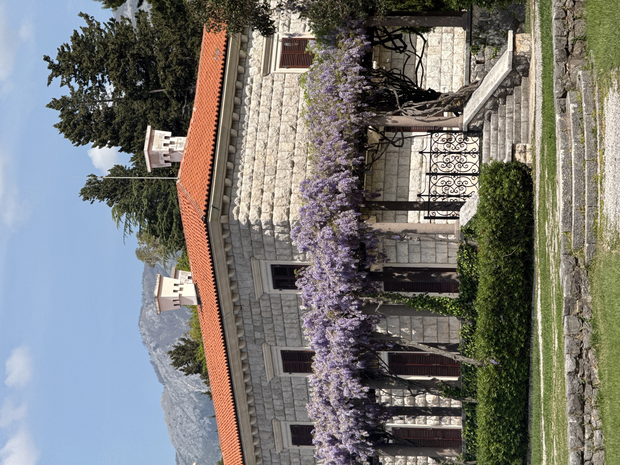 The image shows a building in Sveti Stefan, Montenegro. The building has a brick facade and a red tile roof. A flowering vine grows along the building's facade. The windows have dark shutters. There are stairs with a stone railing to the right of the building. The environment includes a well-kept lawn and lush greenery. In the distance, mountains and blue skies can be seen.