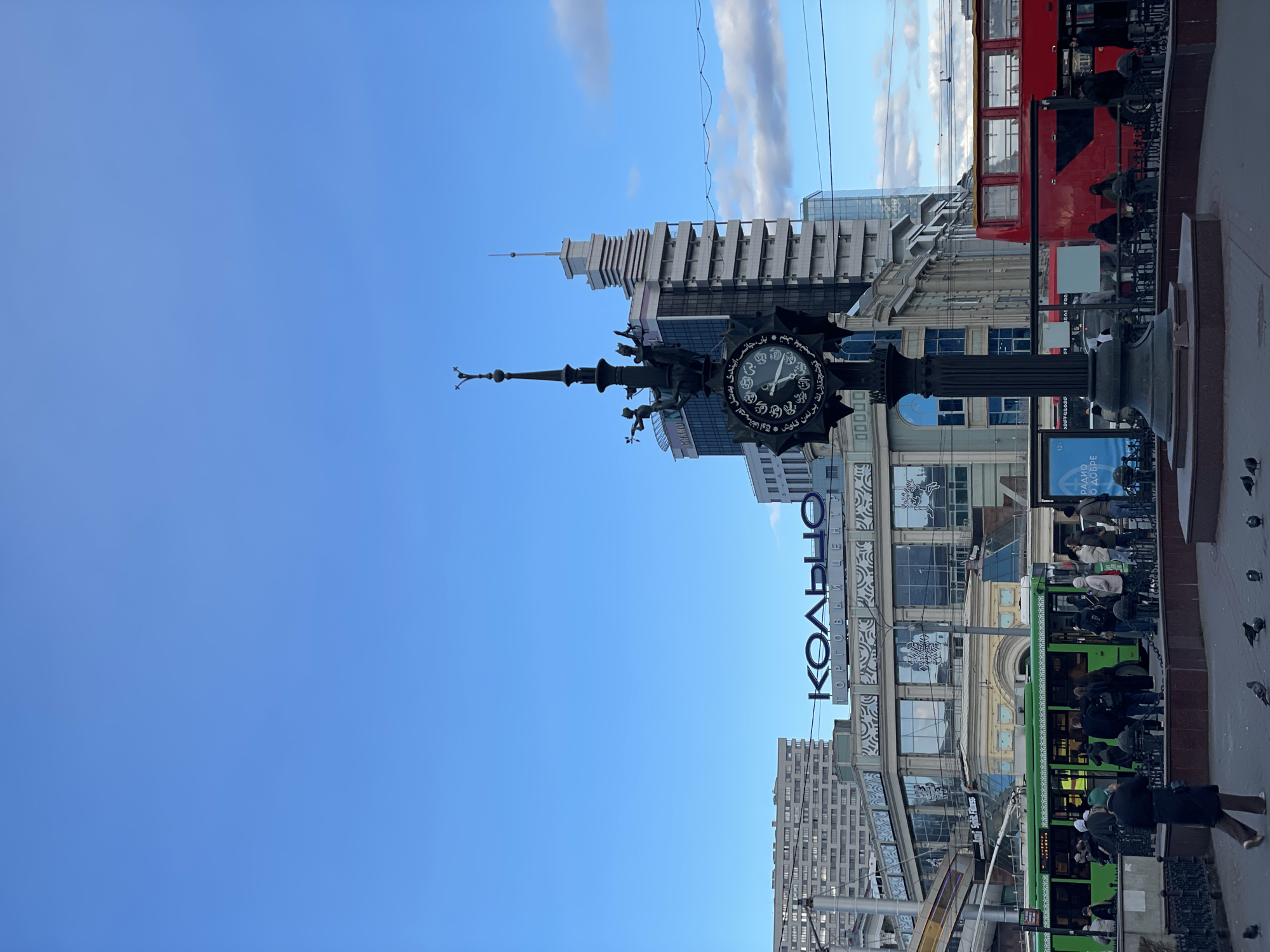 This media file captures a vibrant daytime street scene in Kazan, Russia. The image, viewed upright, showcases a bustling urban environment under a clear blue sky with sparse clouds.
In the foreground, a prominent, ornate street clock stands on a dark pillar. Its circular face features intricate Arabic script, likely displaying Tatar proverbs, and traditional clock hands indicating the time. To the left of the clock, a modern skyscraper with a distinctive tiered design and a pointed spire dominates the skyline. Other buildings of varying architectural styles, including one with classical elements, line the street.
Numerous people are visible on the sidewalk, walking, waiting, or standing, contributing to the lively atmosphere. Public transportation is a key element of the scene, with a bright red double-decker bus passing by on the street and a green tram stopped or moving slowly near the sidewalk, with passengers visible inside and around it. Pigeons are scattered on the paved ground, adding to the urban feel.
A large, illuminated sign is visible above the tram, displaying the Cyrillic text "КОЛЬЦО ТОРГОВЫЙ ЦЕНТ" which translates to "Koltso Shopping Center". Another smaller blue sign in the lower right corner reads "РАДИО О ДОБРЕ" (Radio About Good) and "12+". The overall impression is that of a dynamic city center, likely a commercial or popular thoroughfare.