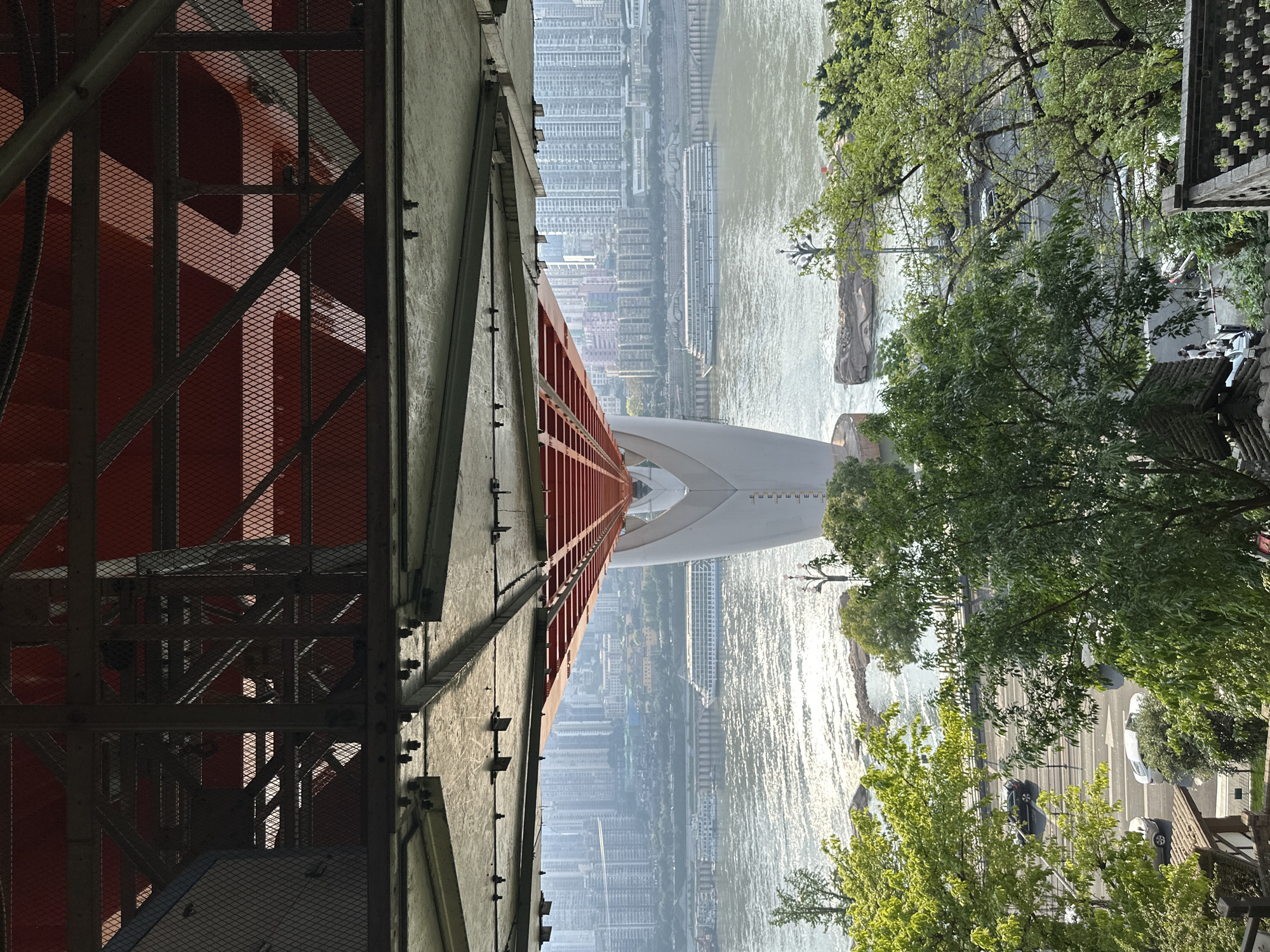 A view of the city from the Nan'an District in China is captured from what appears to be a high vantage point on a bridge structure.
On the left side of the frame is the steel infrastructure of the bridge, painted in red and gray, featuring mesh paneling and cross-bracing. The structure has visible nuts and bolts along the edges.
At the center of the frame, a river is seen with a ship on it. The river reflects sunlight, creating a shimmering effect on the water's surface.
Across the river are buildings. Adjacent to the river is a road with vehicles. The right side of the frame is filled with green foliage.