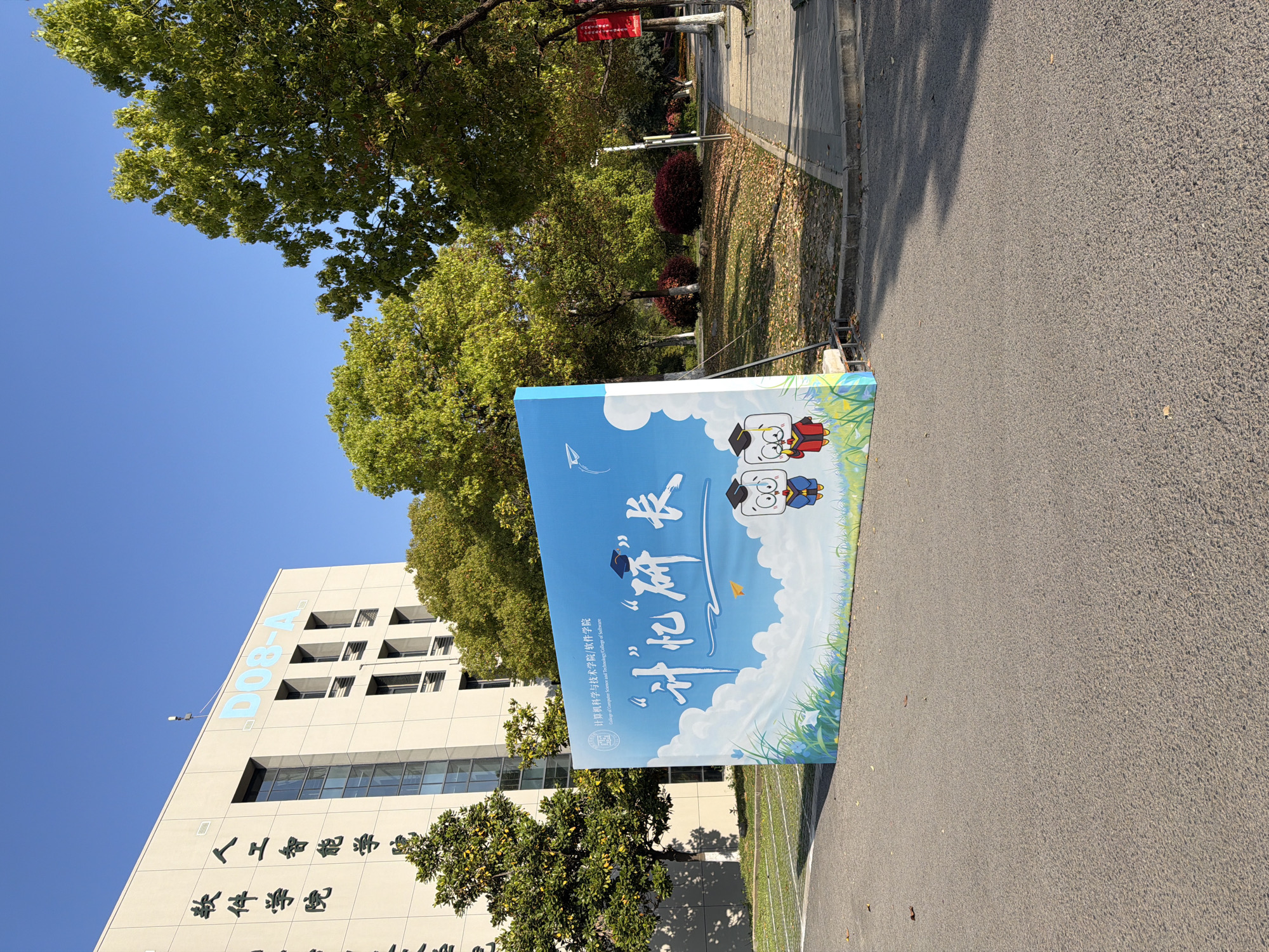 Here is a description of the image:
The image shows an outdoor scene at the College of Computer Science and Technology / College of Software in the Jiangning District of Nanjing, China. The building, labeled "D08-A" with Chinese characters below it, is visible in the background. A large, light-blue banner stands to the right. It features cartoon graduates, clouds, and the Chinese characters 计忆研长, which appear to be part of a promotional message. The banner also includes the English text "College of Computer Science and Technology / College of Software" and the college's logo. The ground is paved, and there are trees and other greenery around. The sky is clear and blue.
